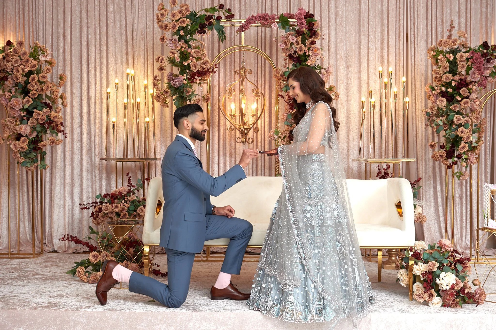 A couple is participating in a wedding ceremony, with the groom kneeling on one knee presenting a ring to the bride who is standing. The setting is decorated with pink and cream flowers, gold accents, and a backdrop of pink curtains, with a sofa and floral arrangements in the background.