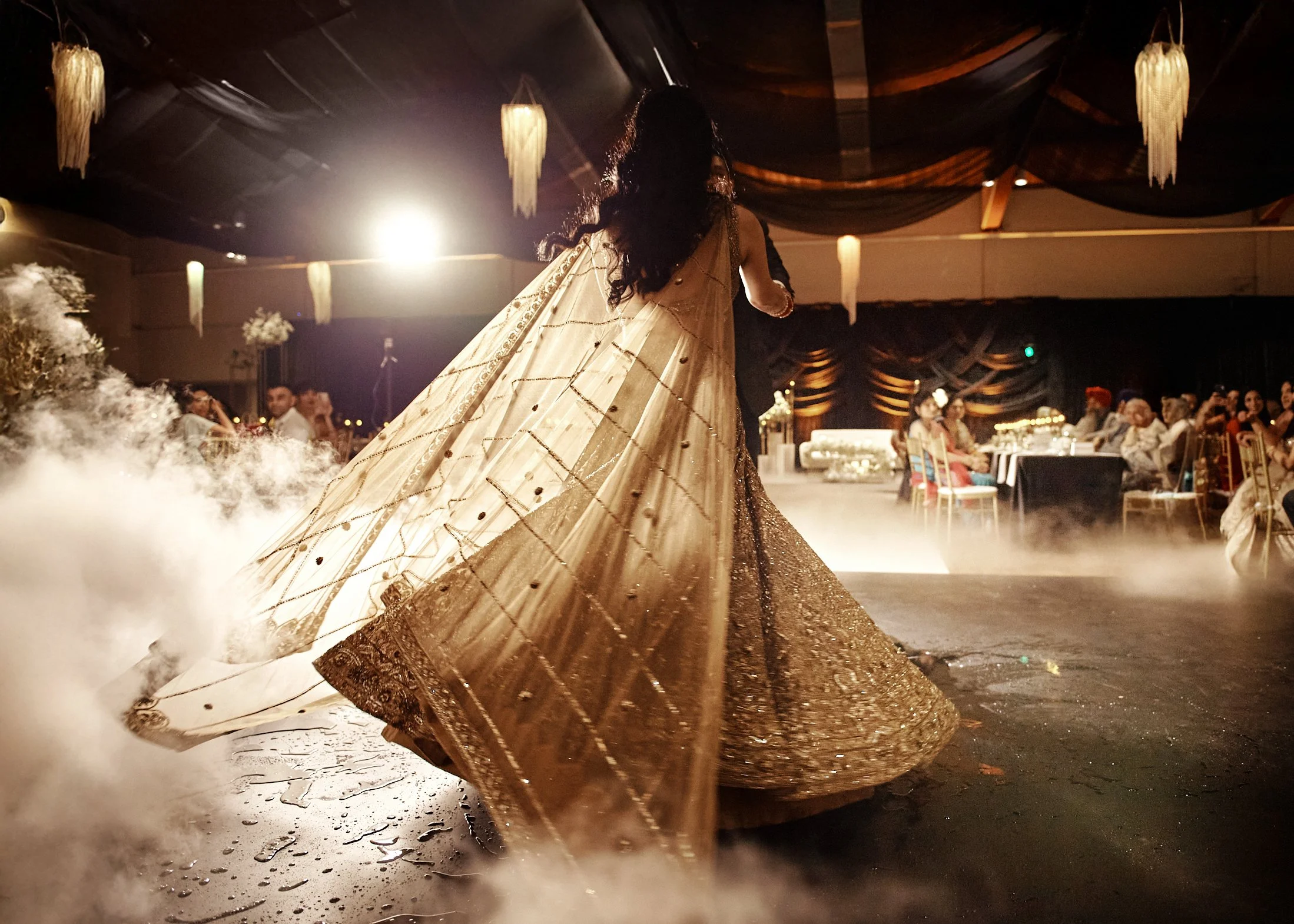 A woman in traditional Indian attire dancing at a wedding reception amid fog and warm lighting, with guests seated at tables in the background.