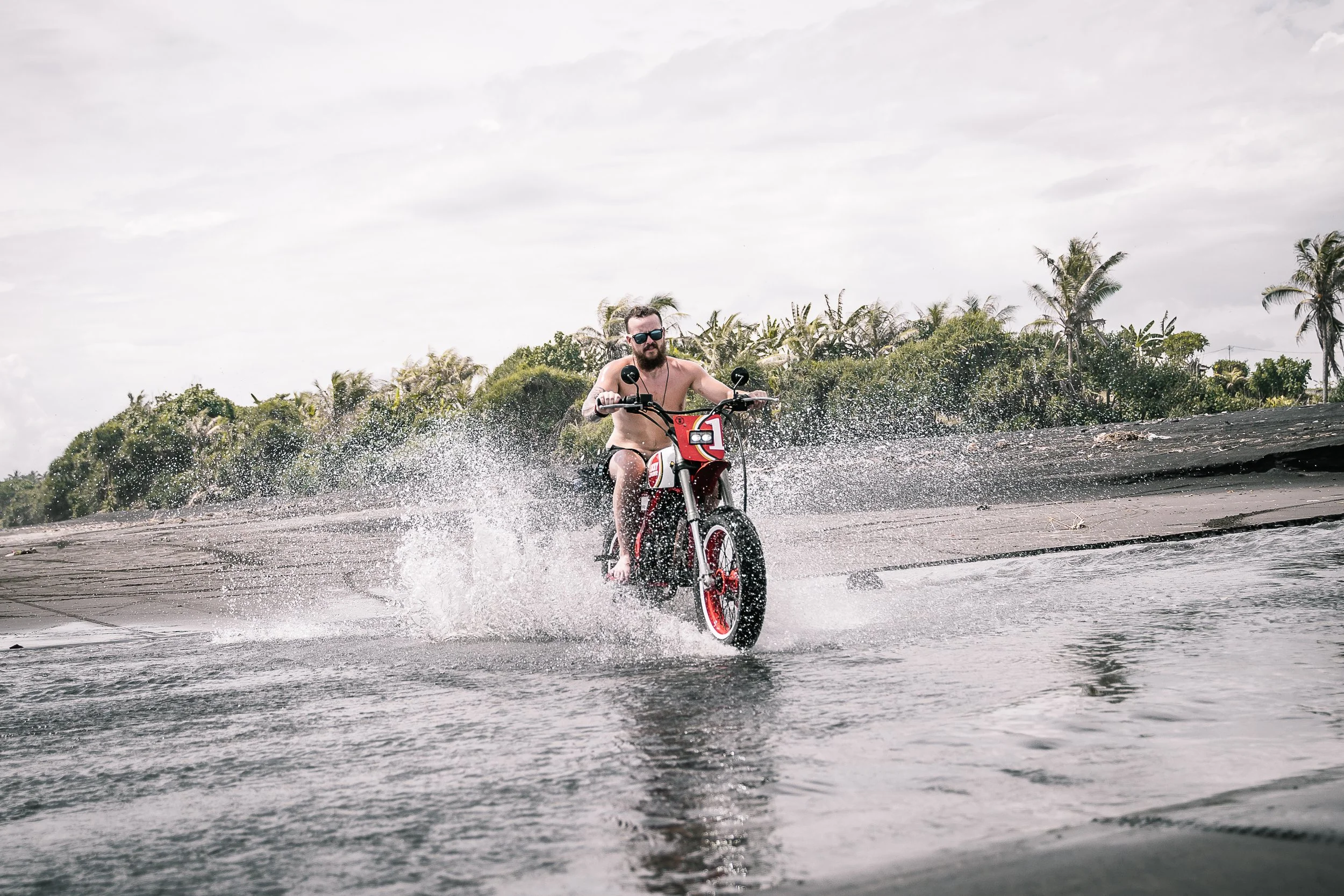 Shirtless man rides a rid and white motorbike through the water with jungle behind him | Edmonton motorcycle photographer DarkShark Media