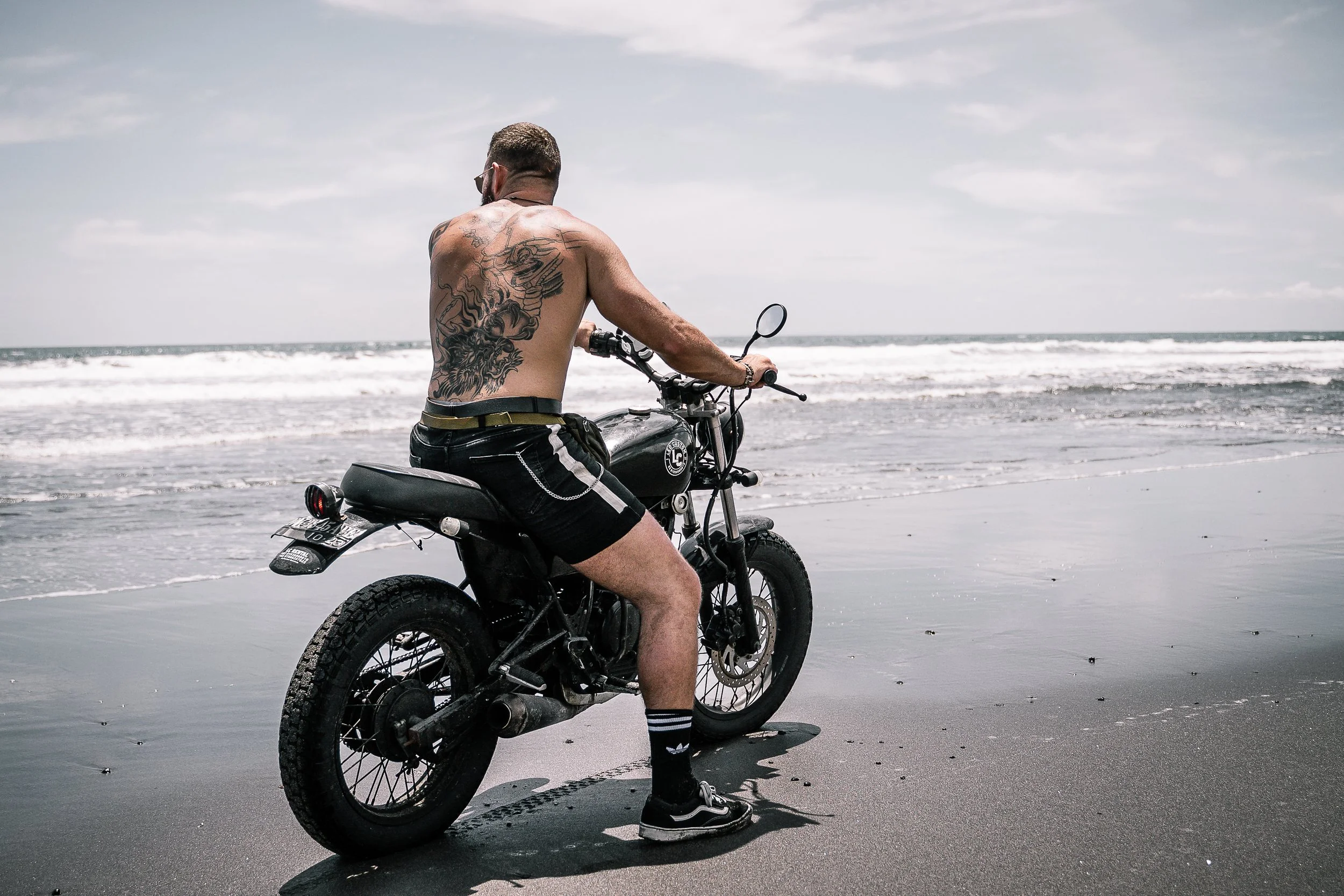 Tattooed man without shirt sits on black motorcycle on beach while looking out at the ocean in Bali, Indonesia | Edmonton motorcycle photographer DarkShark Media