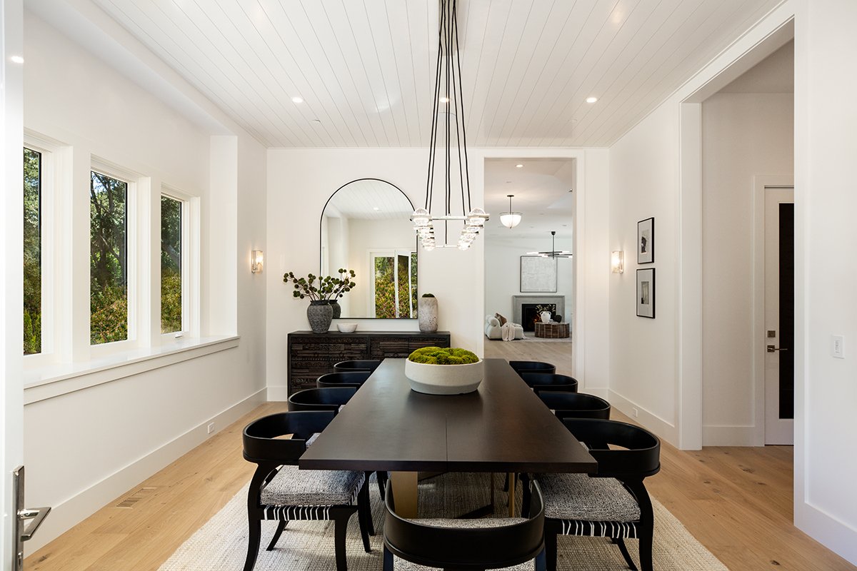 Modern dining room with a long black table, eight black chairs with woven seats, white walls, windows, and a decorative bowl with moss in the center of the table. A large mirror is on the back wall, and there is a sideboard with vases and decorative items. The room has a light wooden floor and a minimalist, contemporary style.