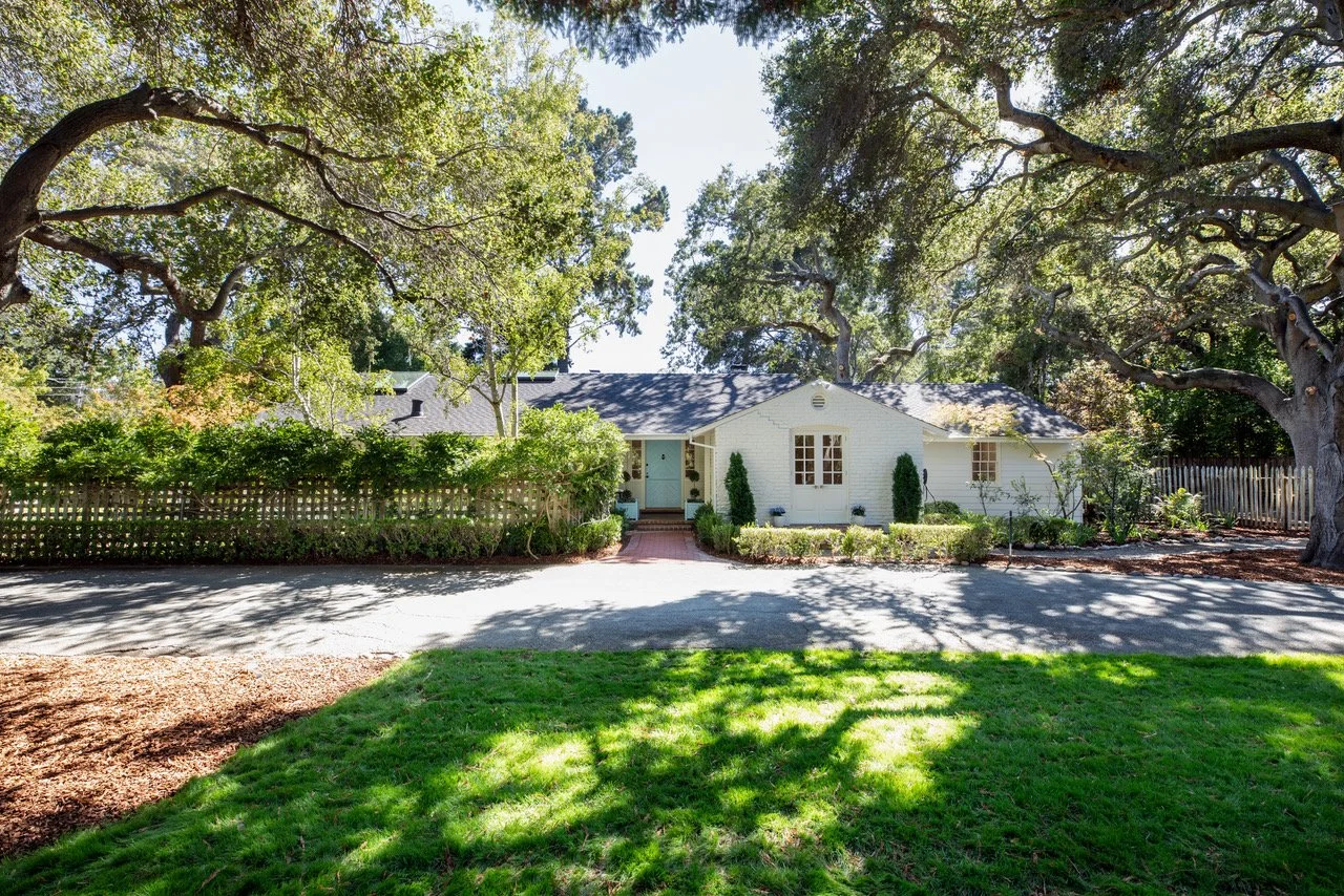 White house with a dark roof surrounded by trees and a lawn, viewed across a driveway with shaded grass in the foreground.