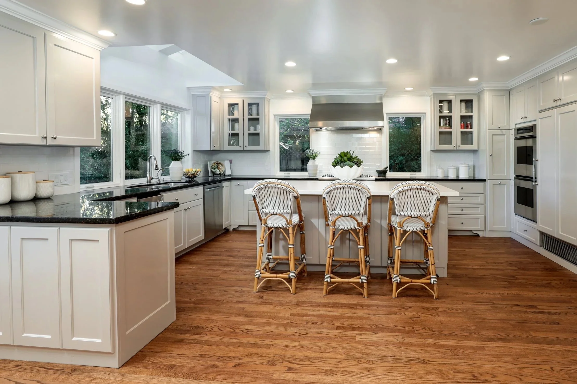 A bright, modern kitchen with white cabinets, black countertops, and hardwood floors. There are three wicker barstools at a kitchen island, stainless steel appliances, and large windows bringing in natural light. Decorative items include potted plants and bowls.