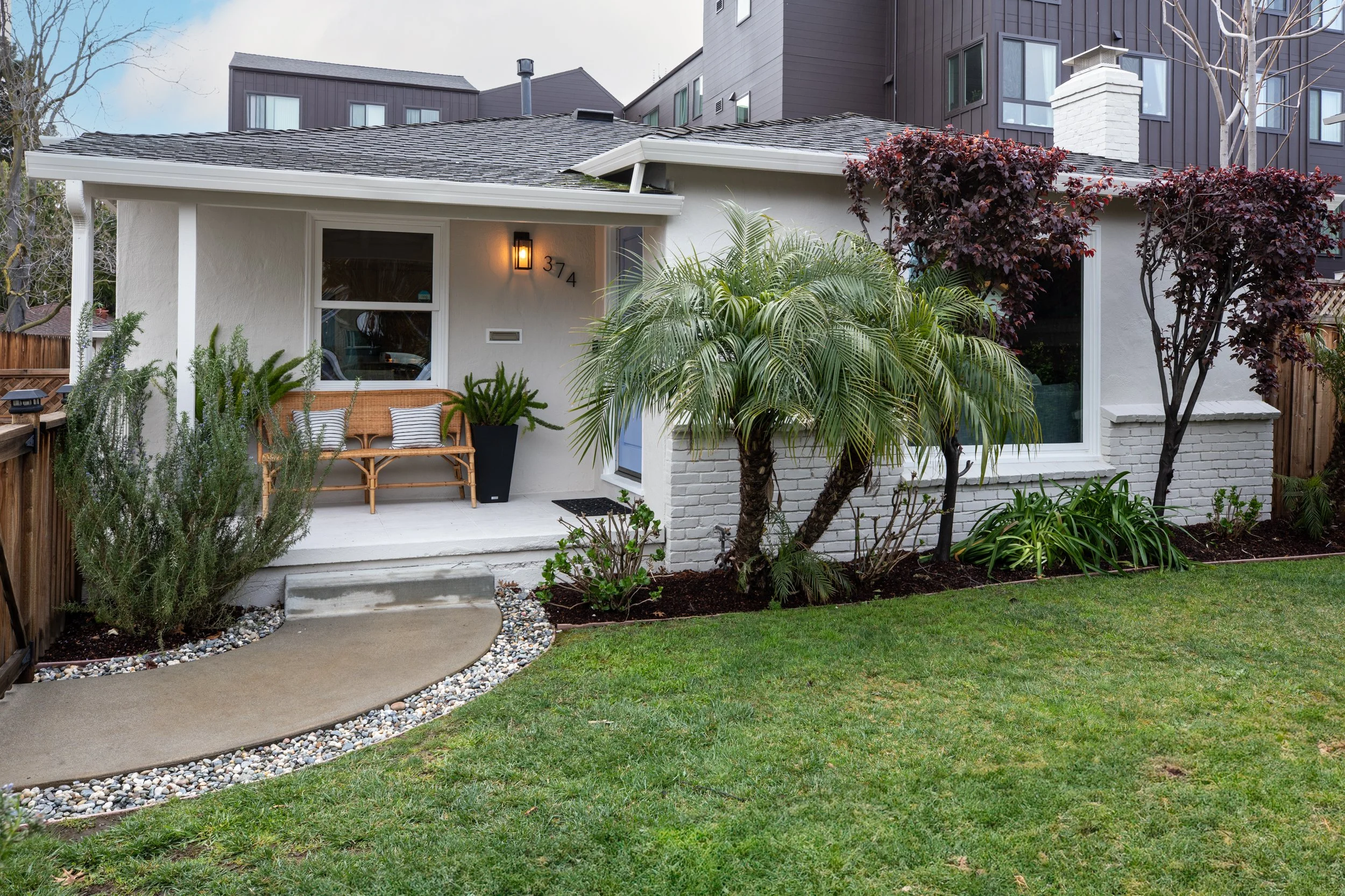 Exterior view of a small white house with a front porch, wooden bench, potted plant, landscaping, trees, and a grassy yard.