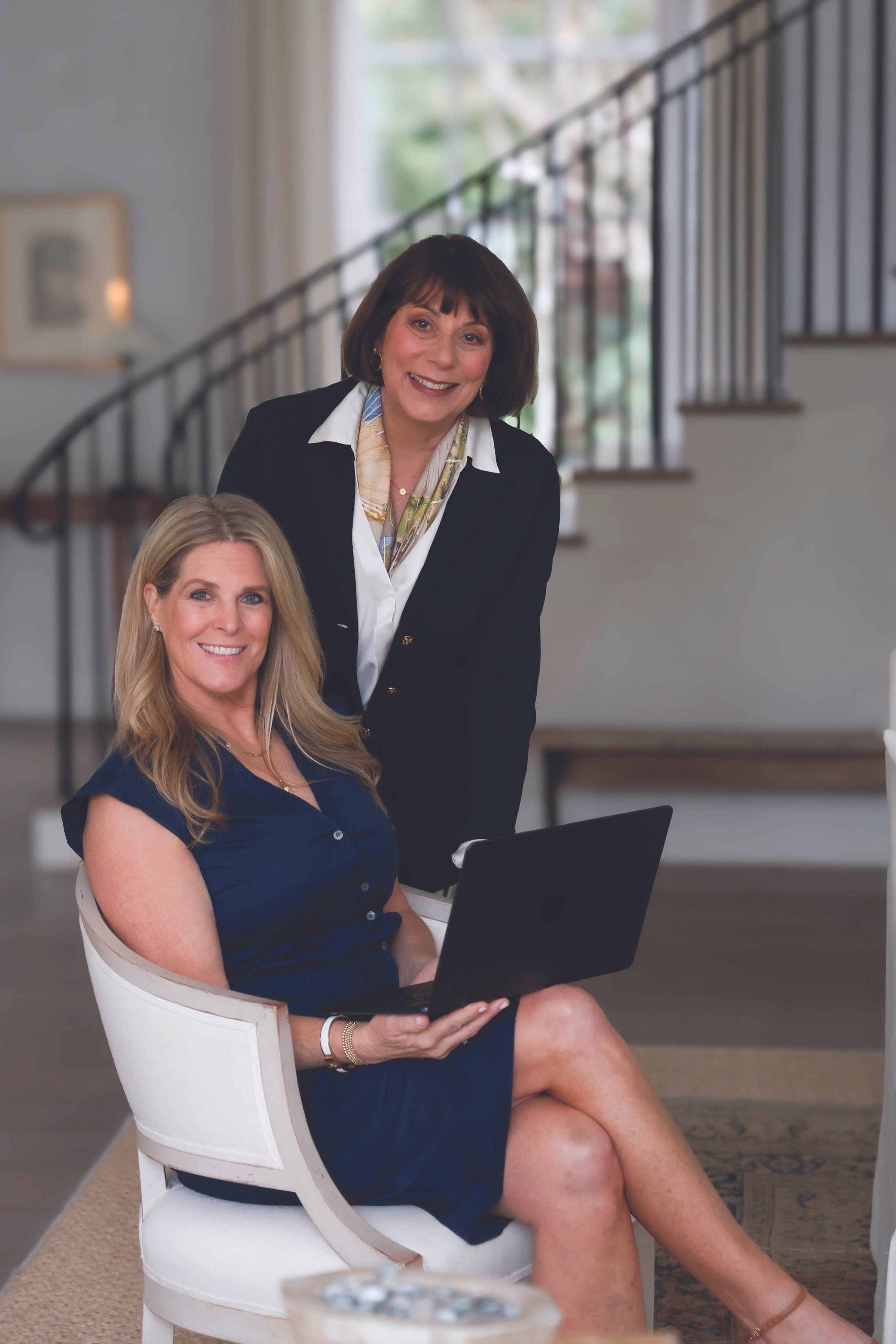 Two women in a home setting, one sitting and using a laptop, the other standing behind her. The woman sitting has blonde hair and is wearing a navy blue dress. The woman standing has dark hair and is wearing a black blazer and a patterned scarf. They are both smiling.