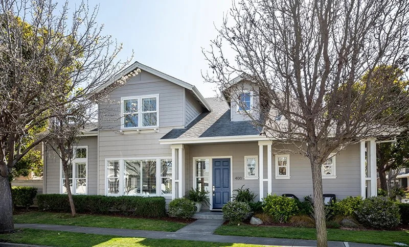 A light gray, two-story house with a blue front door, surrounded by trees and a manicured lawn.