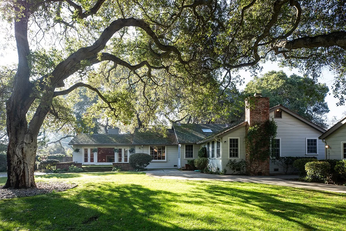 A white house with a brick chimney, surrounded by a large shaded yard with green grass and trees.