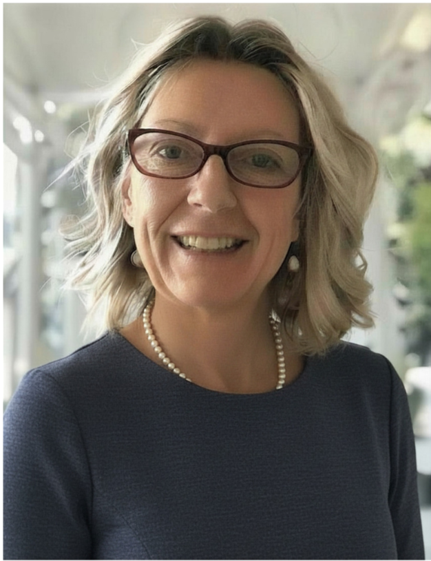 A woman with wavy blonde hair, wearing glasses, pearl earrings, a pearl necklace, and a navy blue top, smiling indoors with a blurred natural background.