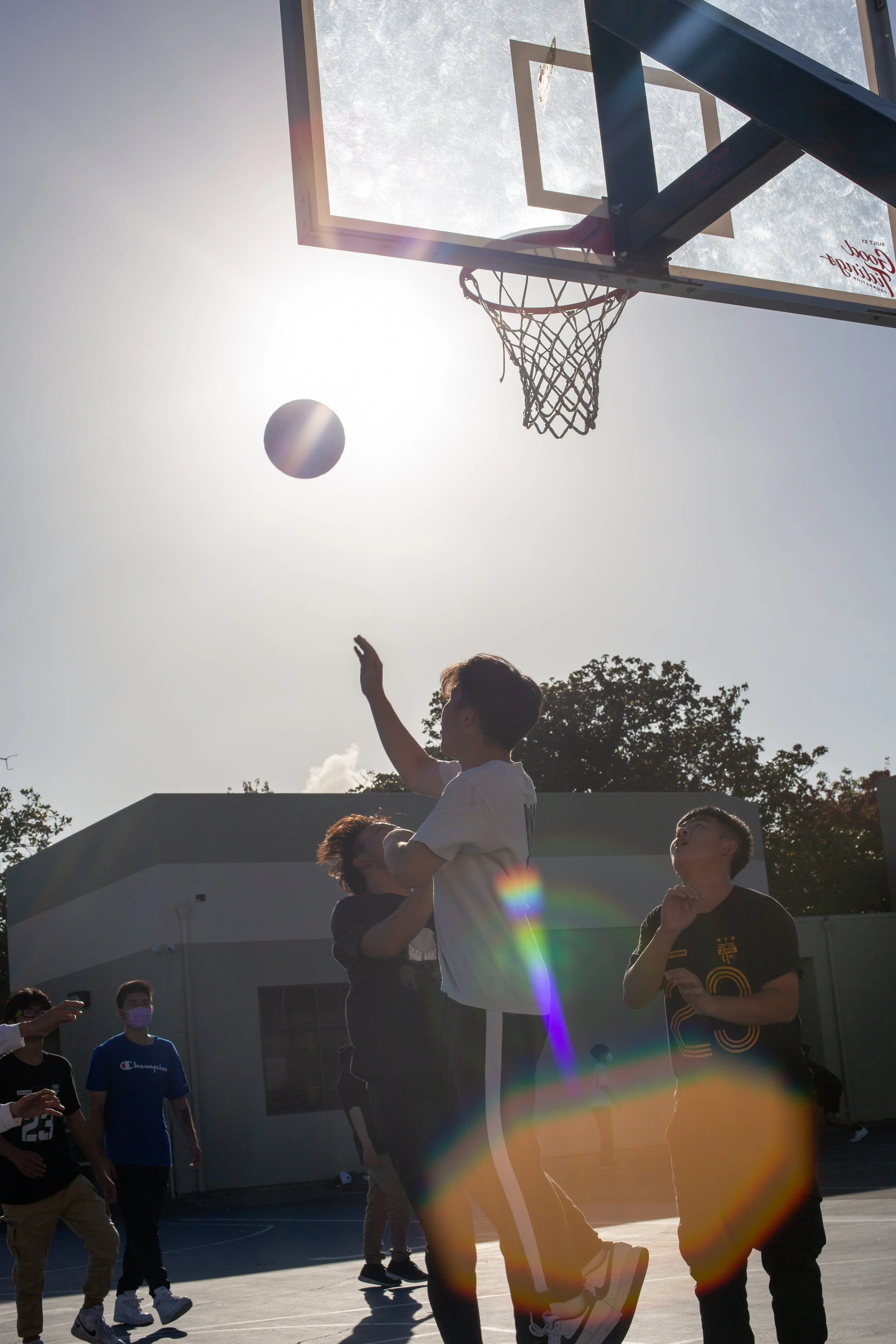 People playing basketball outdoors on a court with a hoop, with the sun shining brightly in the sky and a rainbow lens flare in the foreground. Lincoln Park, Oakland, California. Chinatown. 