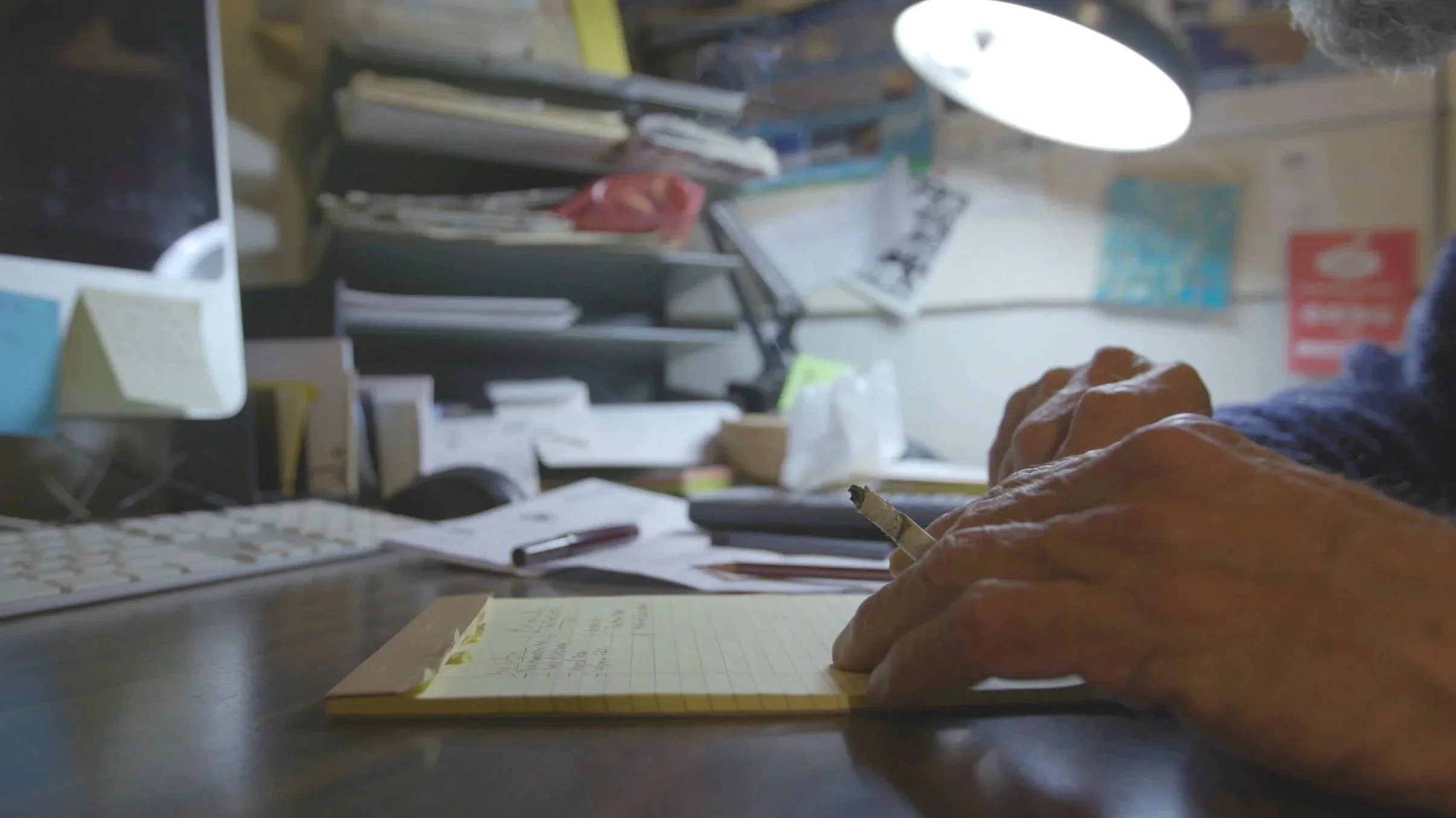 Close-up of elderly person's hands writing on a yellow legal pad at cluttered desk with computer, papers, and office supplies.
