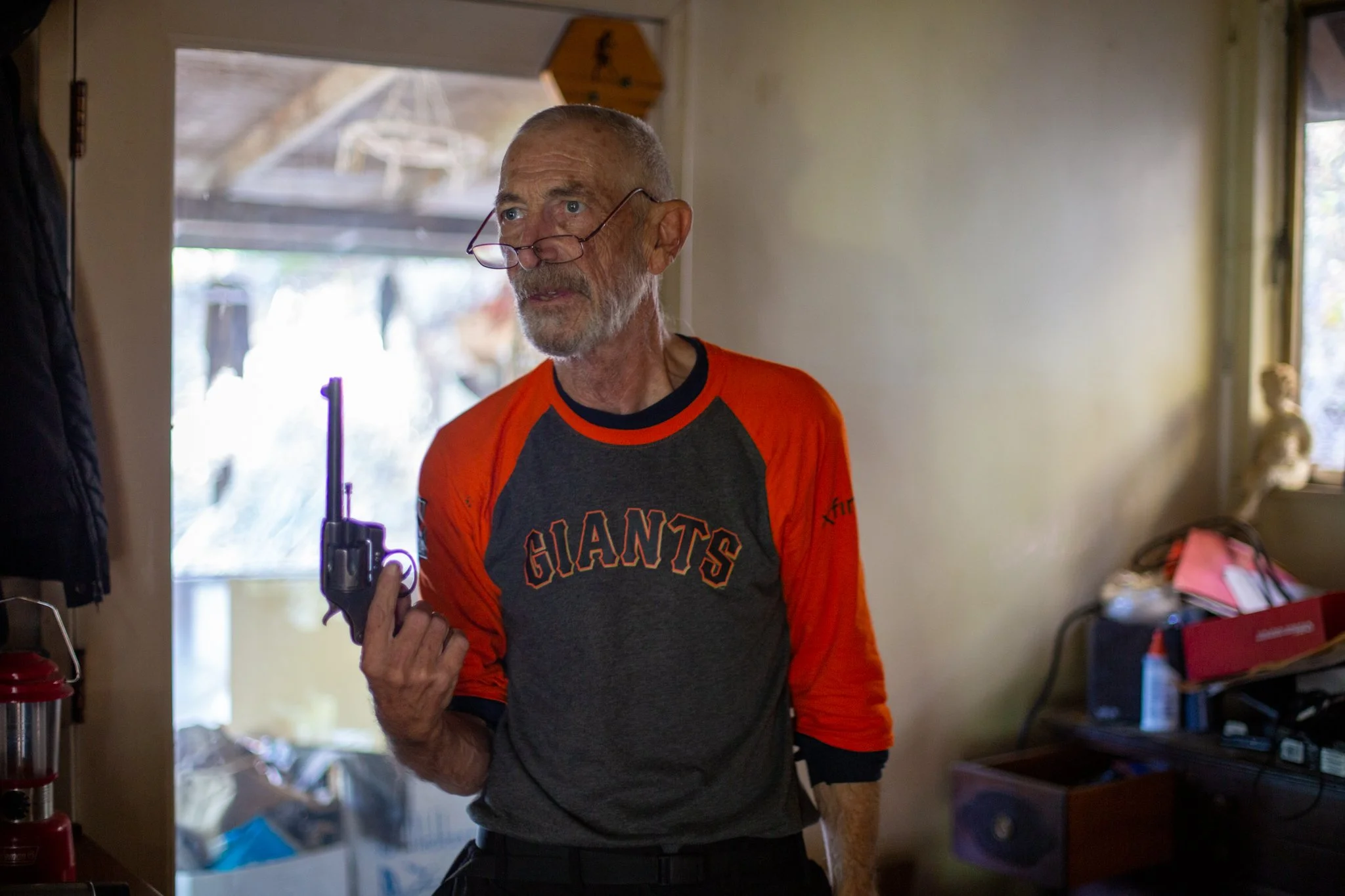 An elderly man (Carl Butz) with glasses, wearing a black and orange Giants sports jersey, holding a small black revolver in his right hand, indoors with cluttered background. From documentary Carl Runs the Paper.  