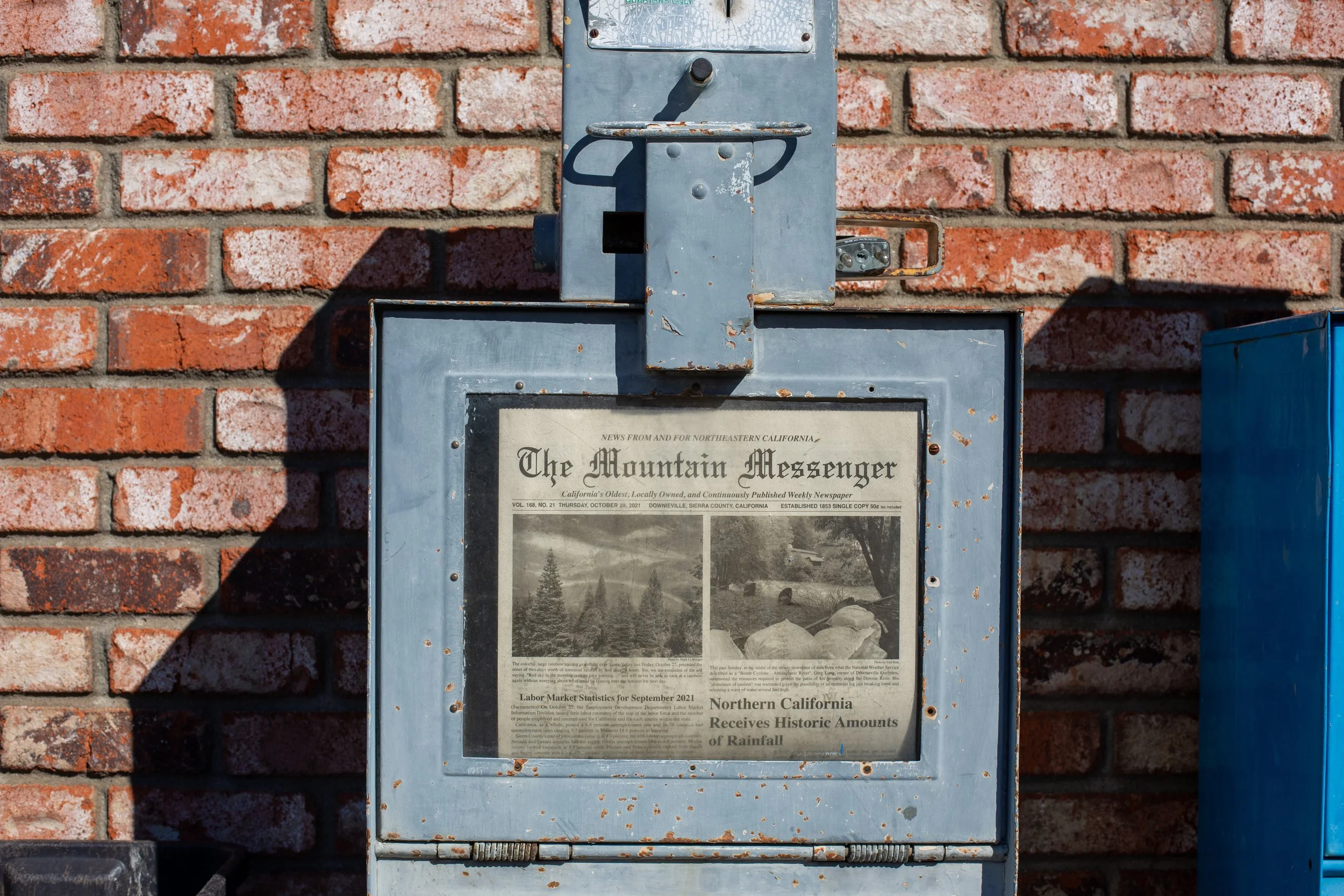 A newspaper box attached to a brick wall containing a copy of 'The Mountain Messenger' newspaper.