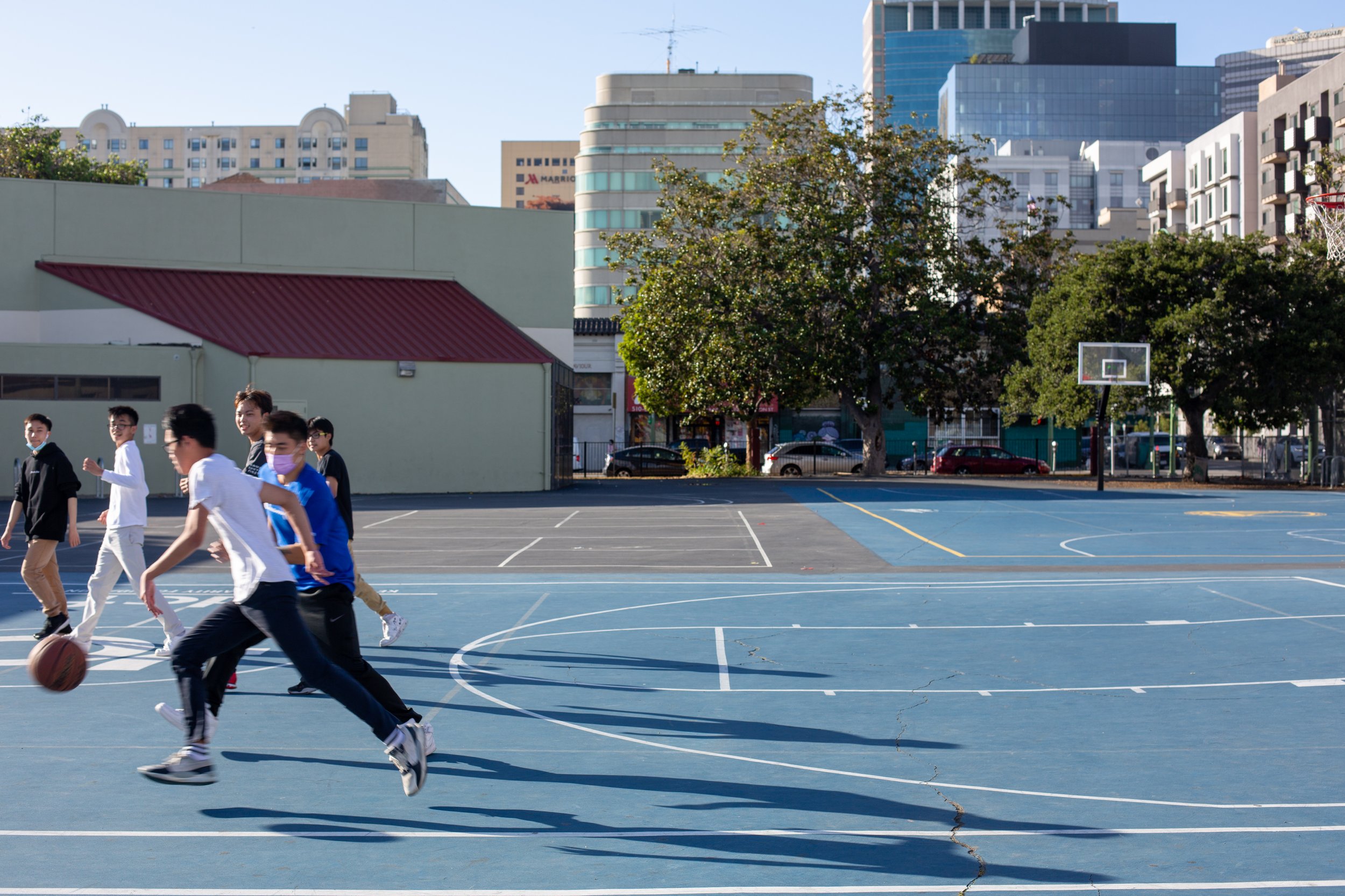 Group of young people playing basketball on an outdoor court with city buildings and trees in the background. Oakland Chinatown. 