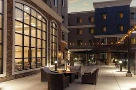 Outdoor seating area with tables and chairs, illuminated by string lights, in front of a building with large windows at dusk.