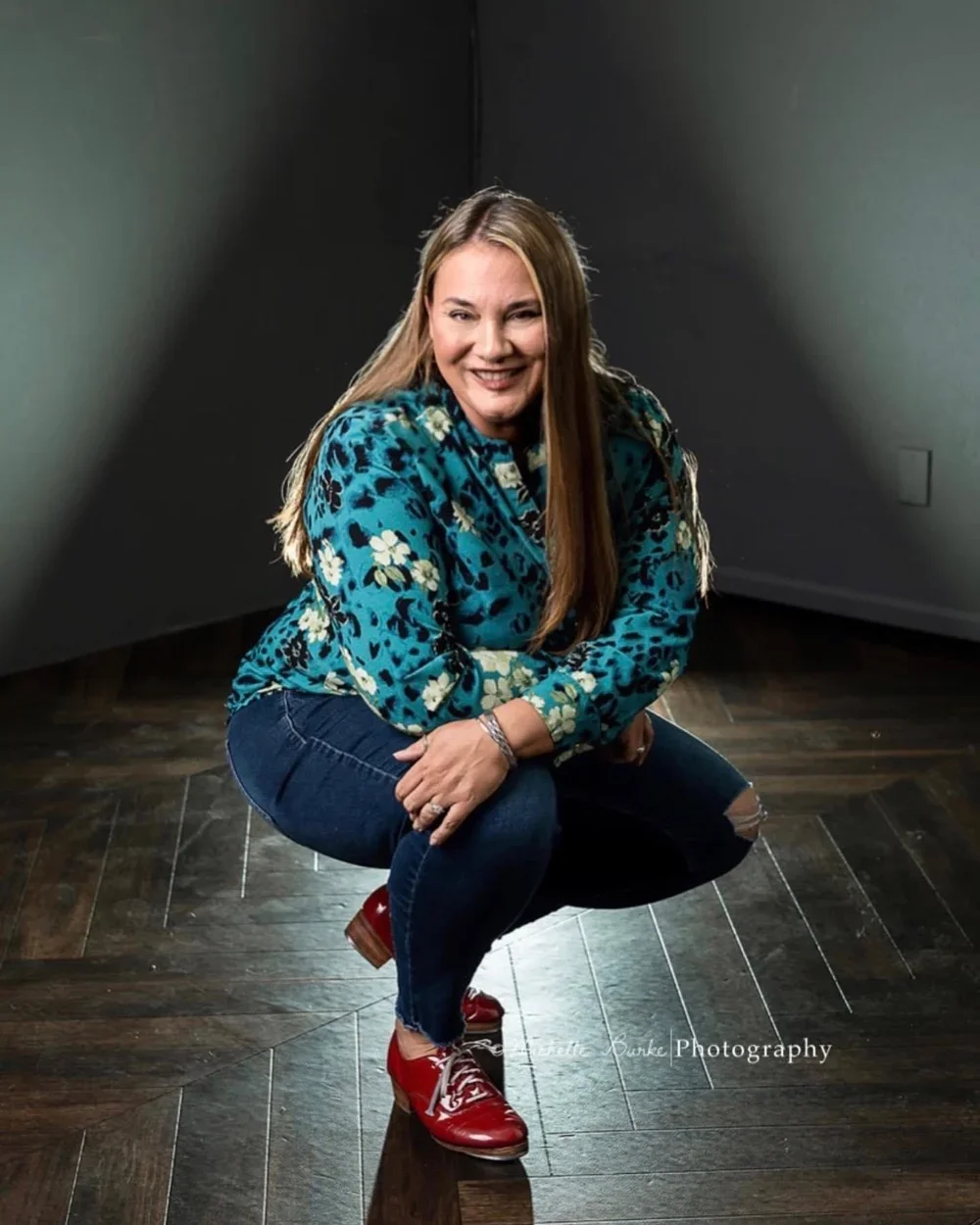 A woman crouching with a smile, wearing a blue floral blouse, dark jeans, and red shoes, in a studio with a dark background and wooden floor.