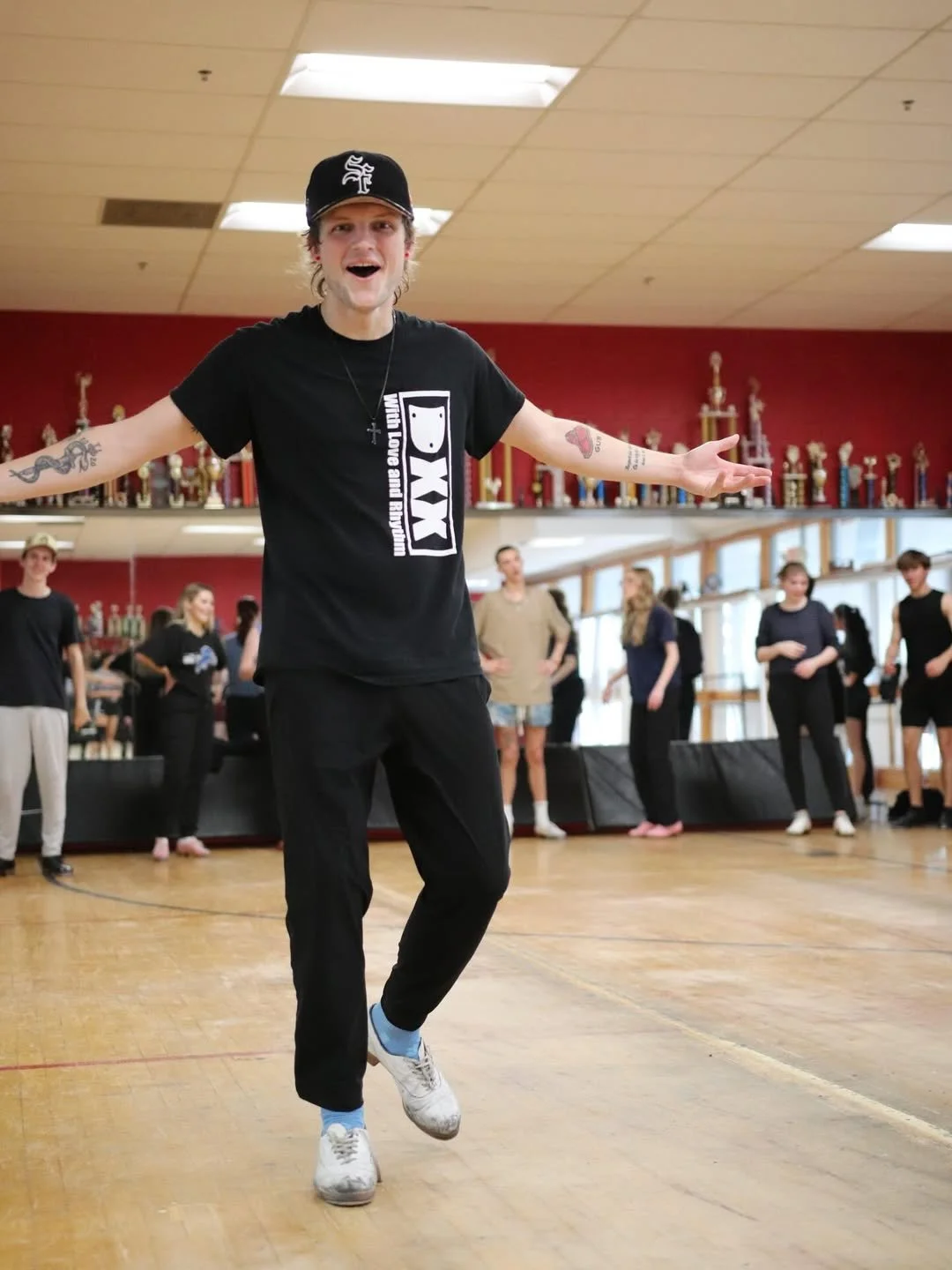 Young man with arms outstretched, wearing a black PXG t-shirt, black pants, white sneakers, and a black cap, standing on a wooden dance floor with other people and trophies in the background.
