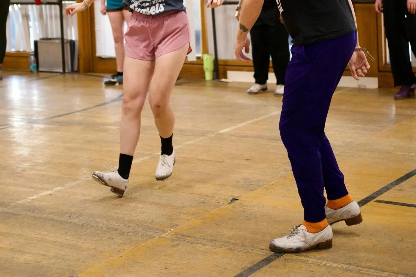 People participating in a dance class, focusing on their legs and feet, on a wooden floor in an indoor studio.