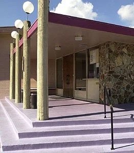 Exterior of a building with stone walls, glass doors, and a porch with steps leading up to it. Wooden posts support the roof overhang.