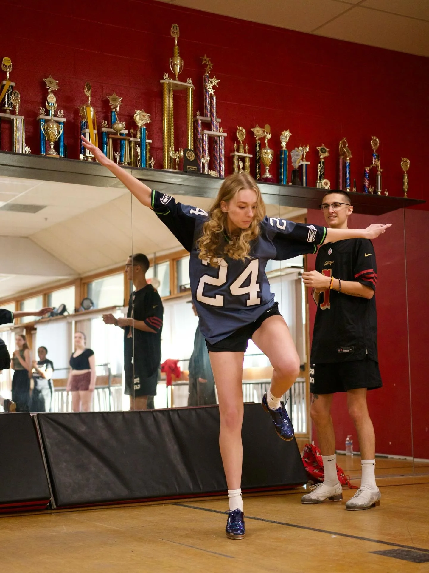 A young woman stretching her arms and balancing on one leg in a dance studio, wearing a sports jersey with the number 24, with a man standing nearby and several trophies on a shelf above them.