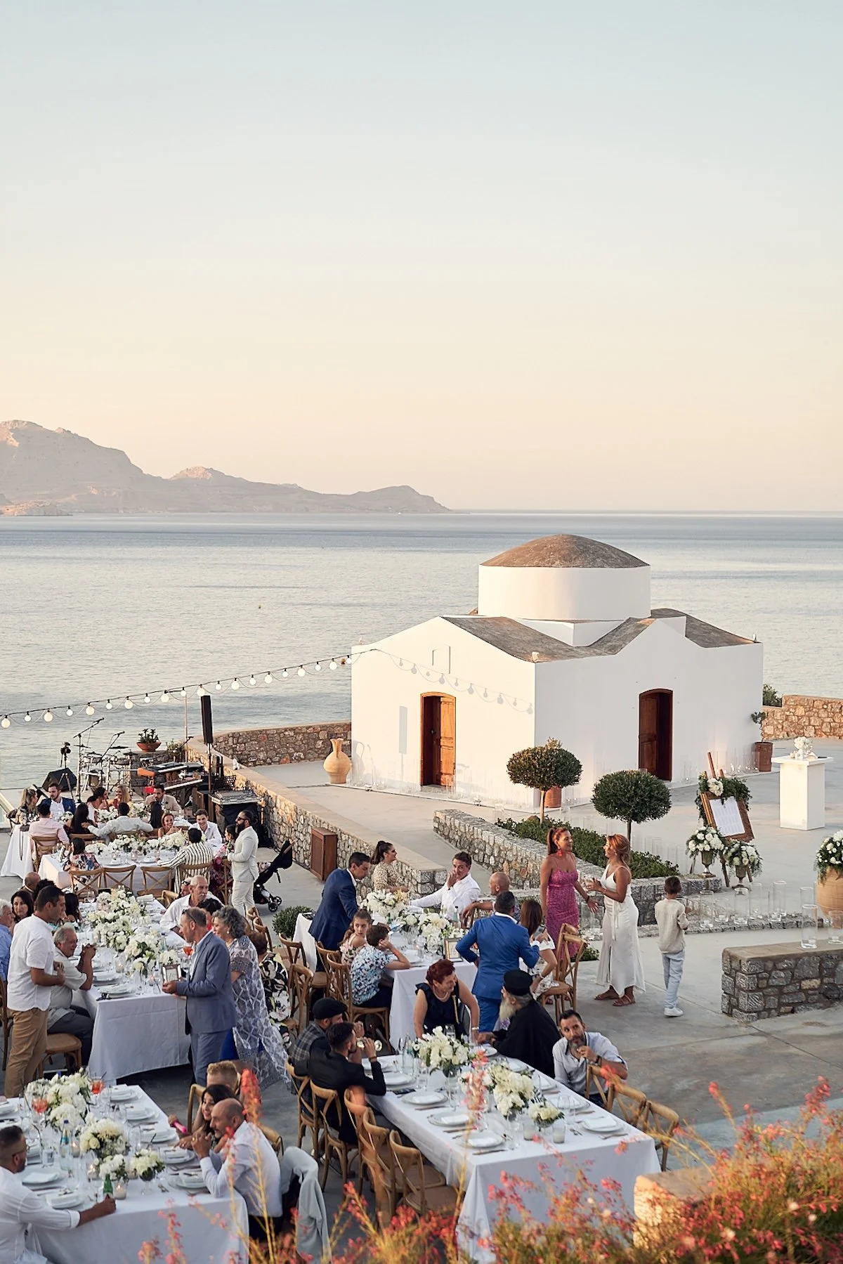 Guests dining al fresco at sunset wedding reception beside a white chapel during a Santorini destination wedding.