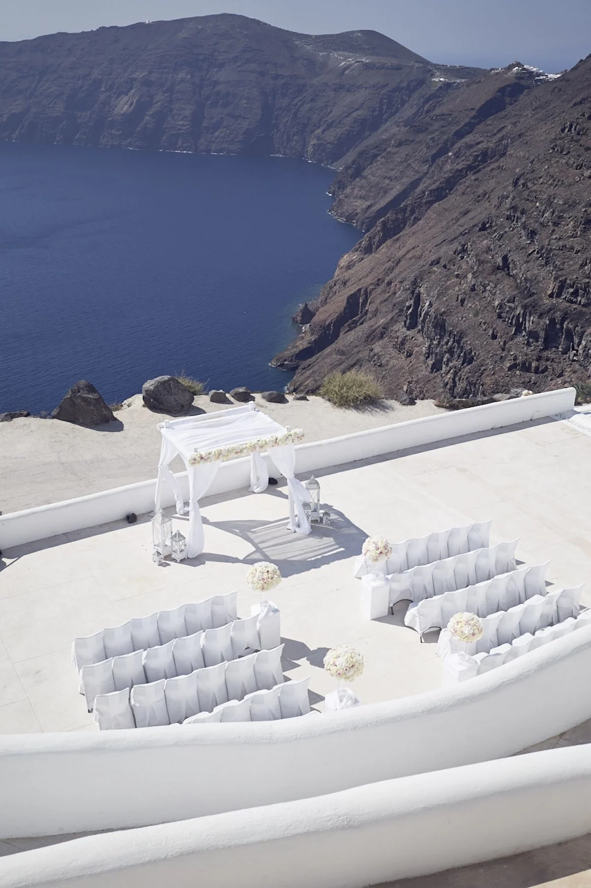 Santorini wedding ceremony setup overlooking the caldera with white chairs, a floral alter structure, and cliffside views.