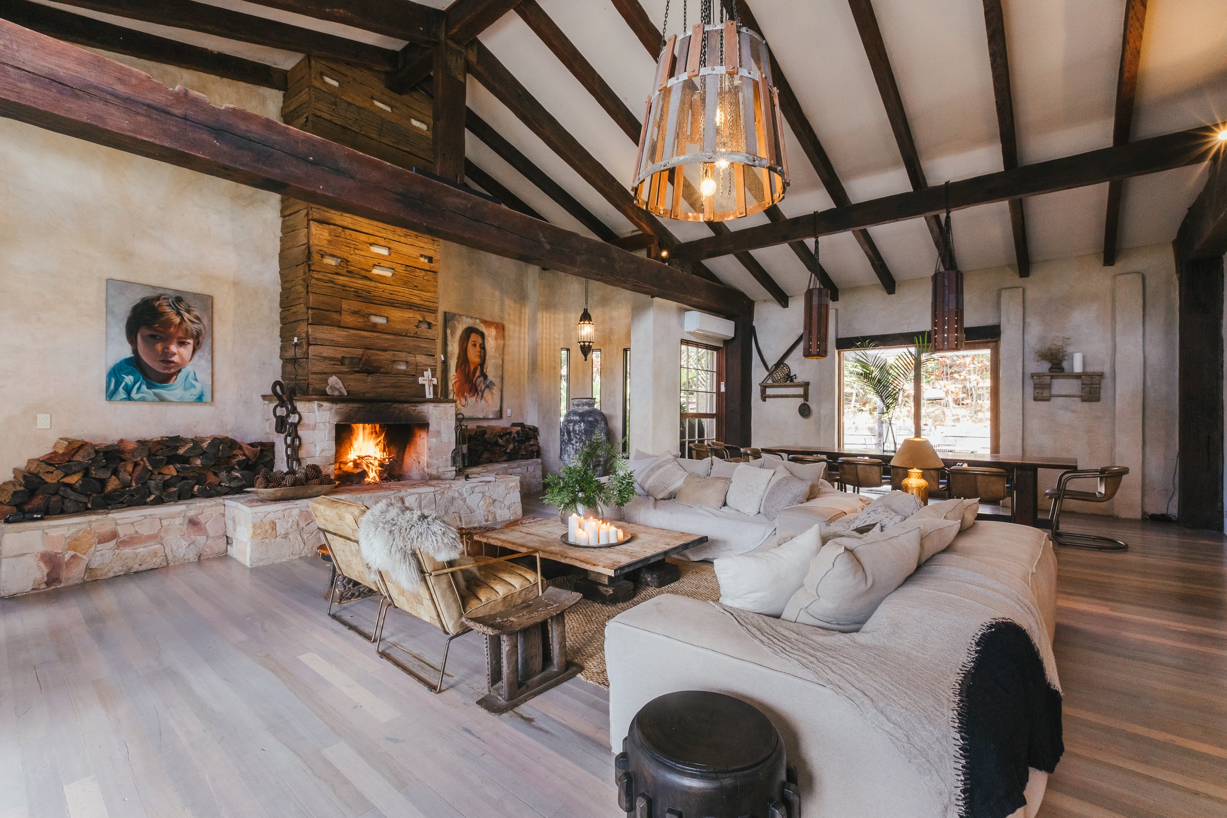 A cozy living room with a fireplace, beige sectional sofa, wooden coffee table with candles, and artwork of children on the walls. There are large windows, pendant lights, and a mix of rustic and modern decor.