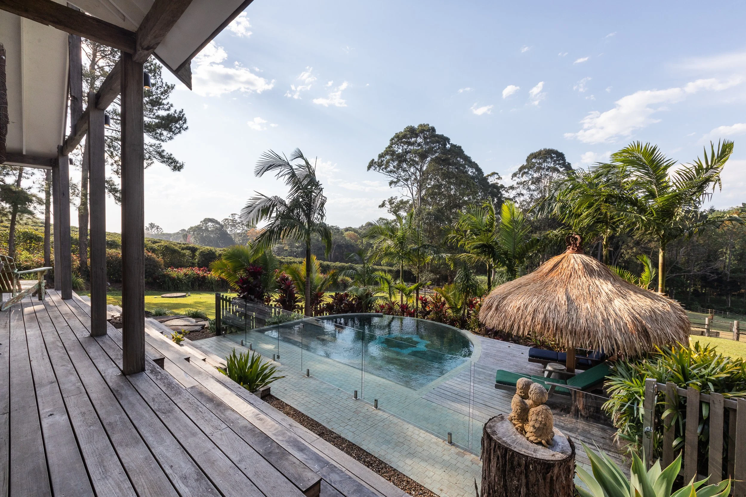 A backyard with a wooden deck overlooking a small pool with a glass barrier. There is a thatched-roof cabana beside the pool, surrounded by tropical plants and trees, under a bright blue sky with some clouds.