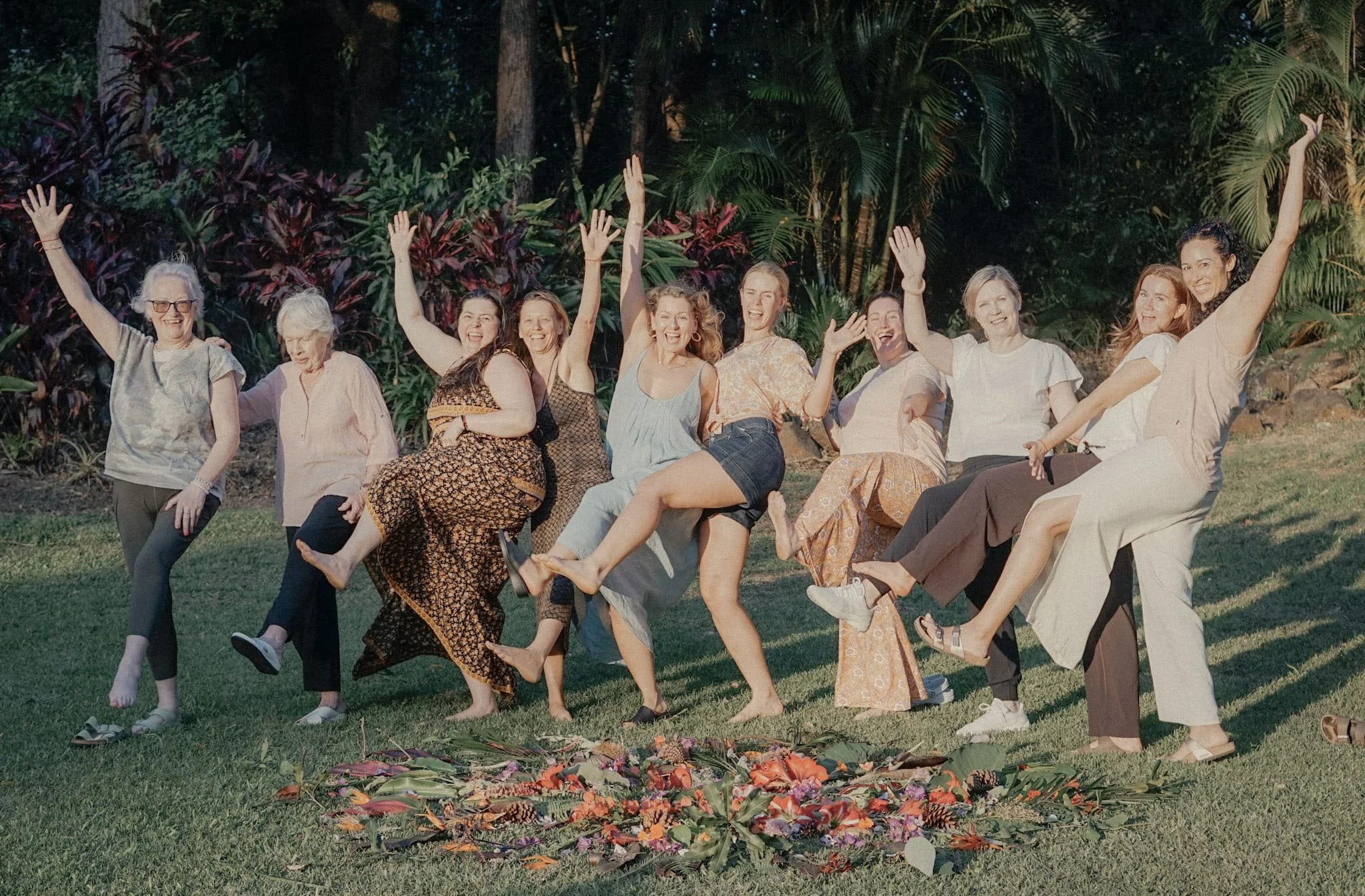 A group of ten women standing outdoors on a grassy area, smiling and raising their arms, with various plants and trees in the background, and colorful flowers and foliage arranged on the ground in front of them.