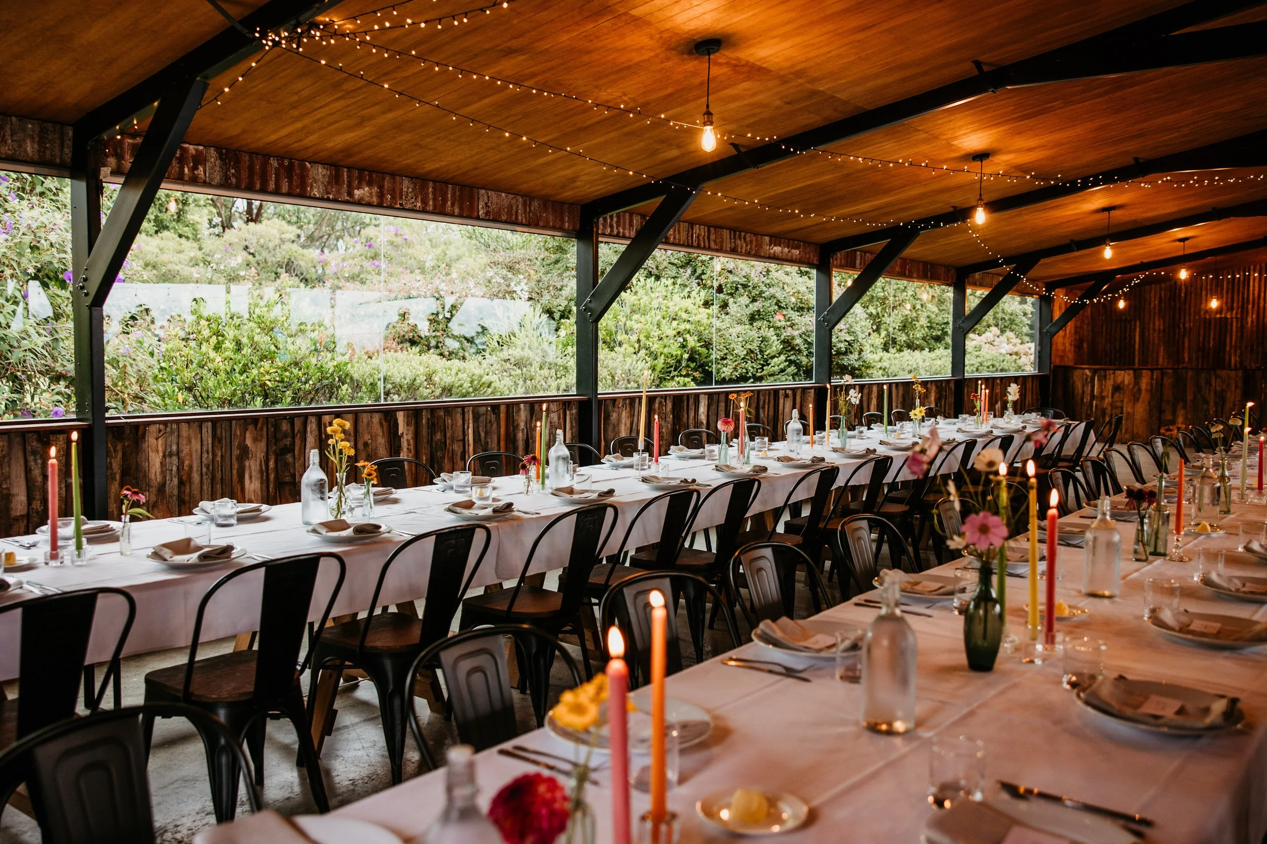 alt = "A grand dining room set for a wedding celebration with white table cloths, fine cutlery, stemware and crockery, beautiful floral arrangements in vintage vases, colourful hand made candles and mood lighting."