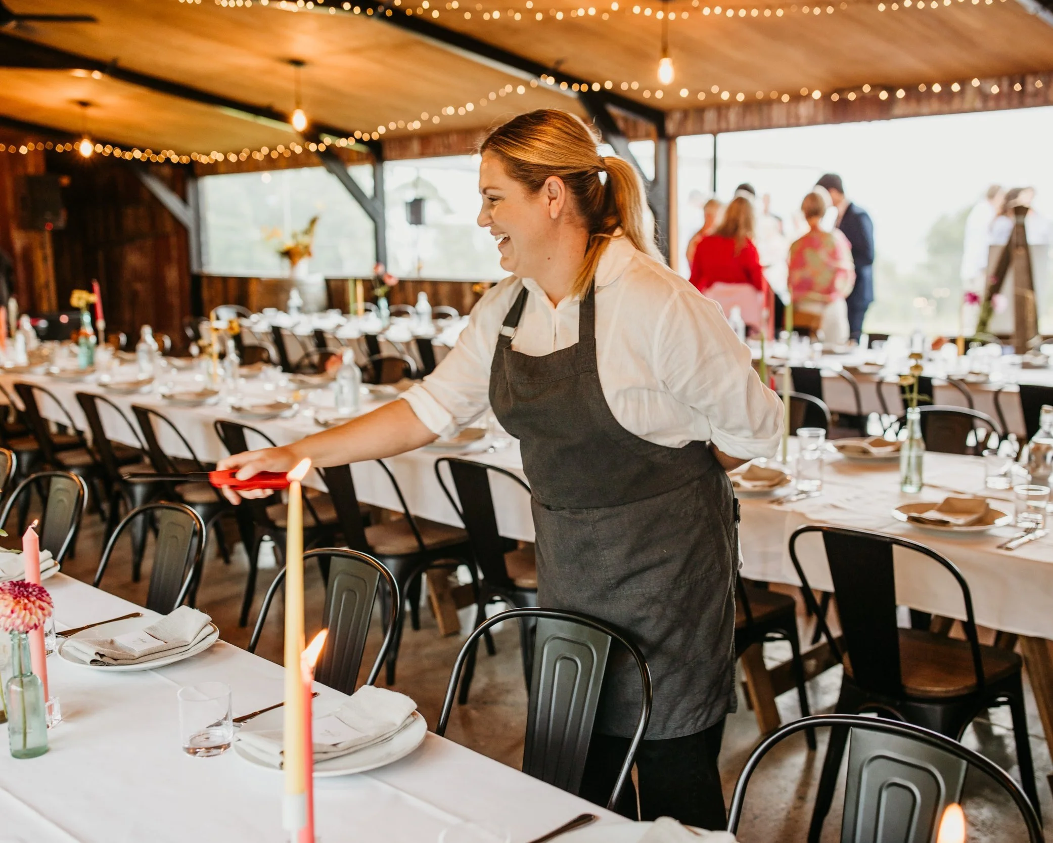 Our talented manager Lucey in a white shirt and black apron is lighting candles on a long banquet table at a rustic event space with string lights and large windows.