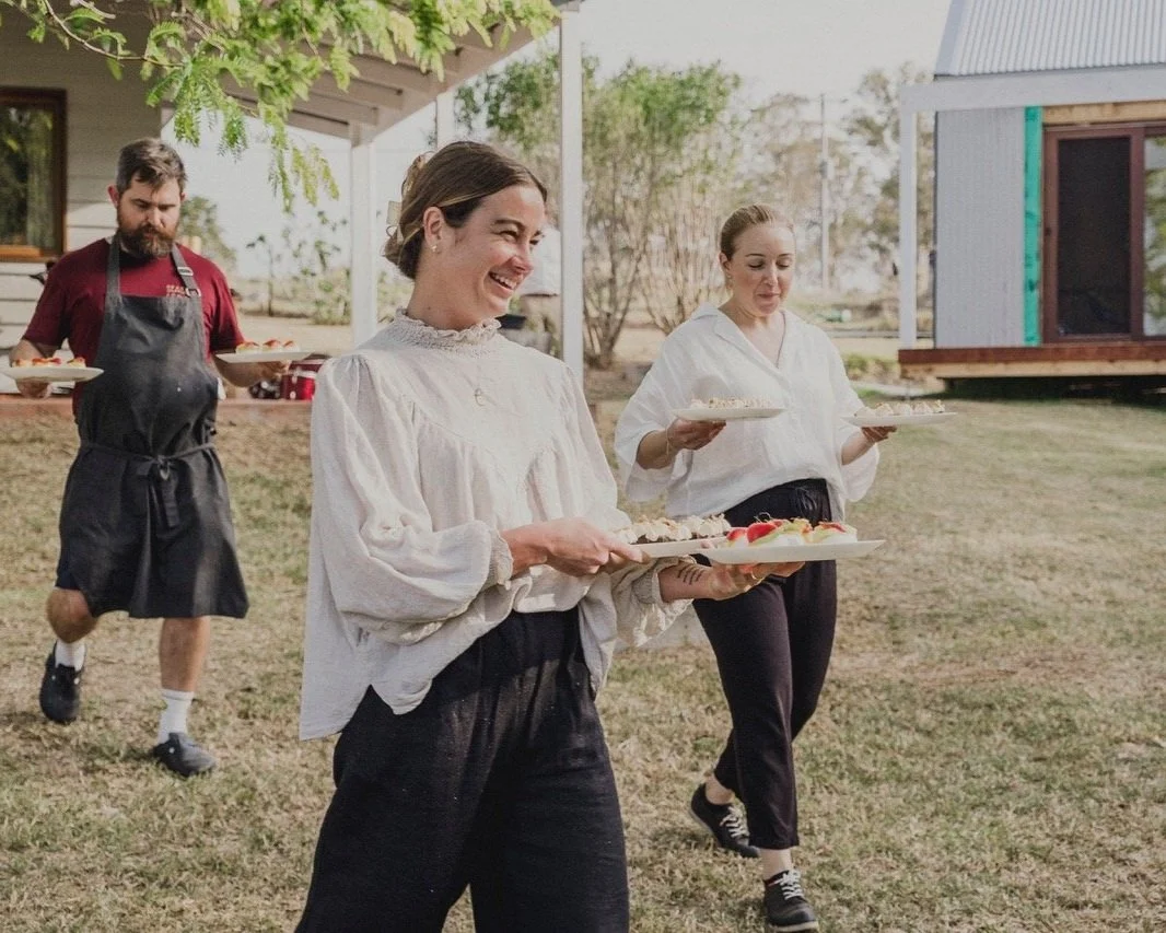 Three of the Season & Savour team carrying plates of food outdoors, smiling and serving at a gathering.