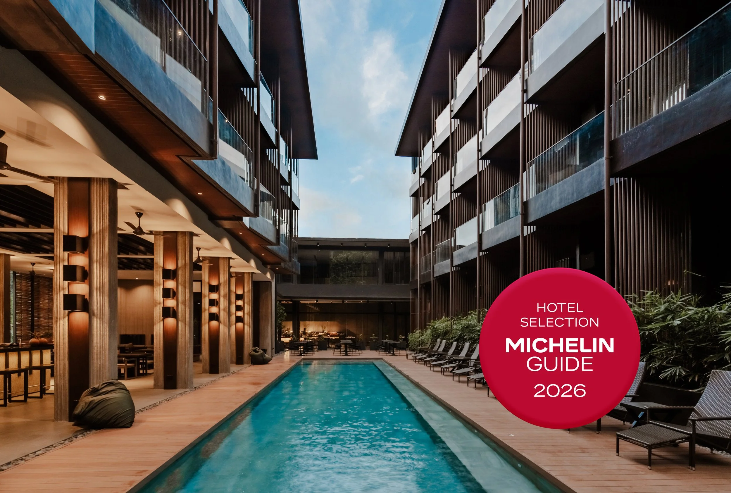 Modern hotel courtyard with a narrow swimming pool, surrounded by lounge chairs and greenery, with multiple balconies and a glass facade.
