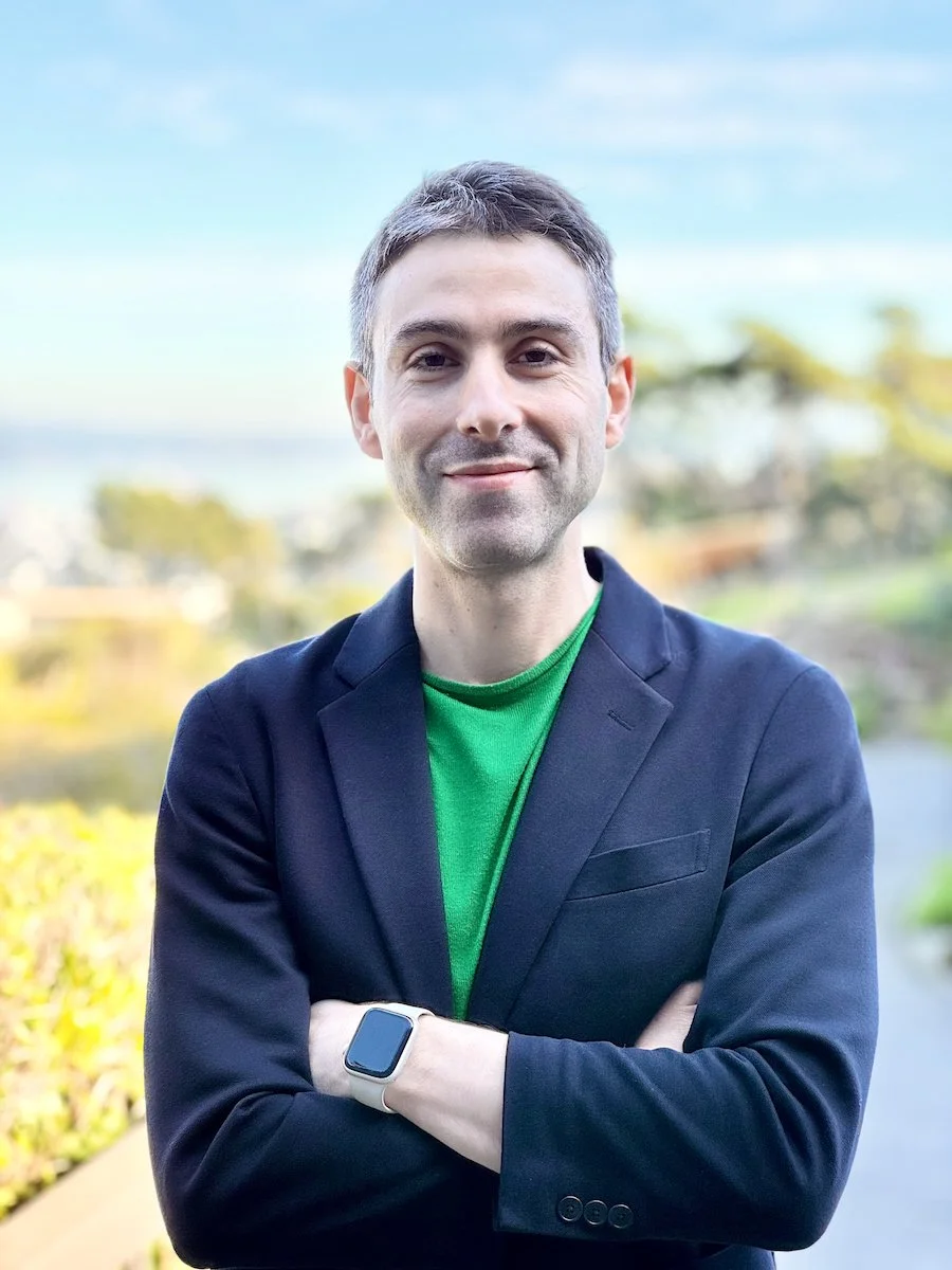 A young man with short brown hair, dressed in a dark blazer and green shirt, standing outdoors with arms crossed.