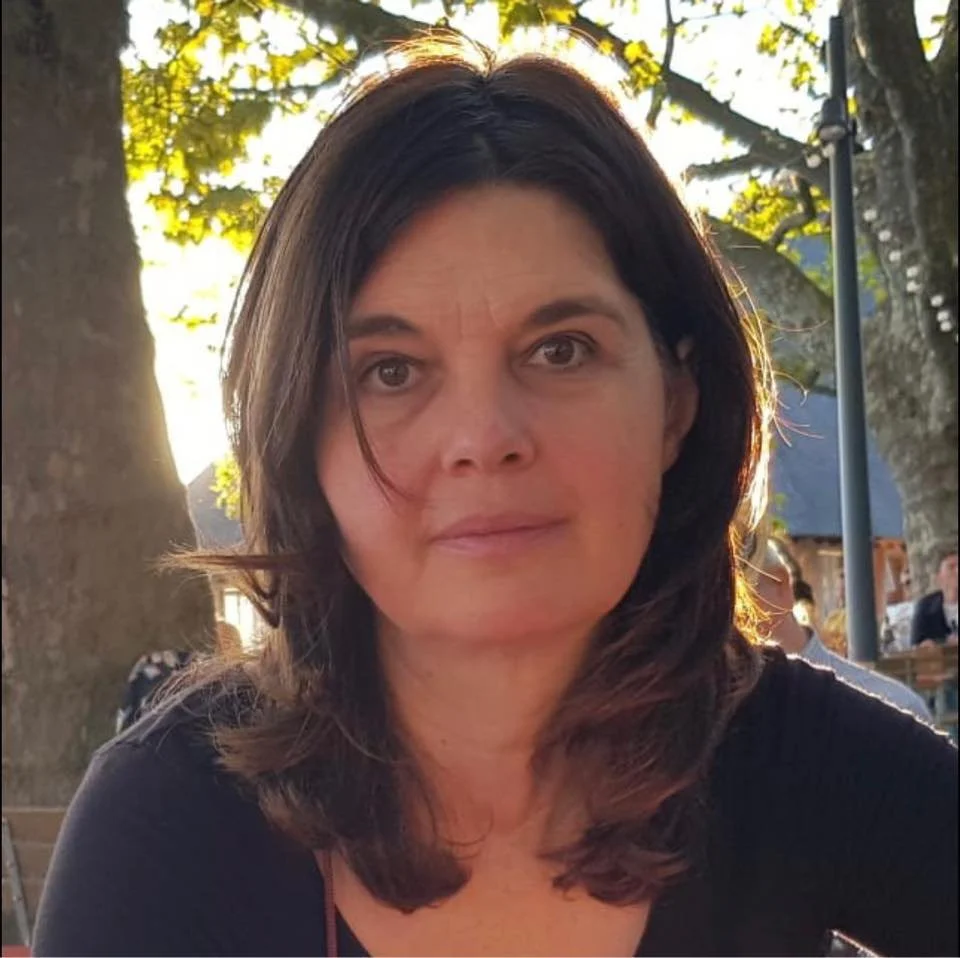 Woman with dark hair sitting outdoors near a large tree, with sunlight filtering through the leaves, and other people in the background.