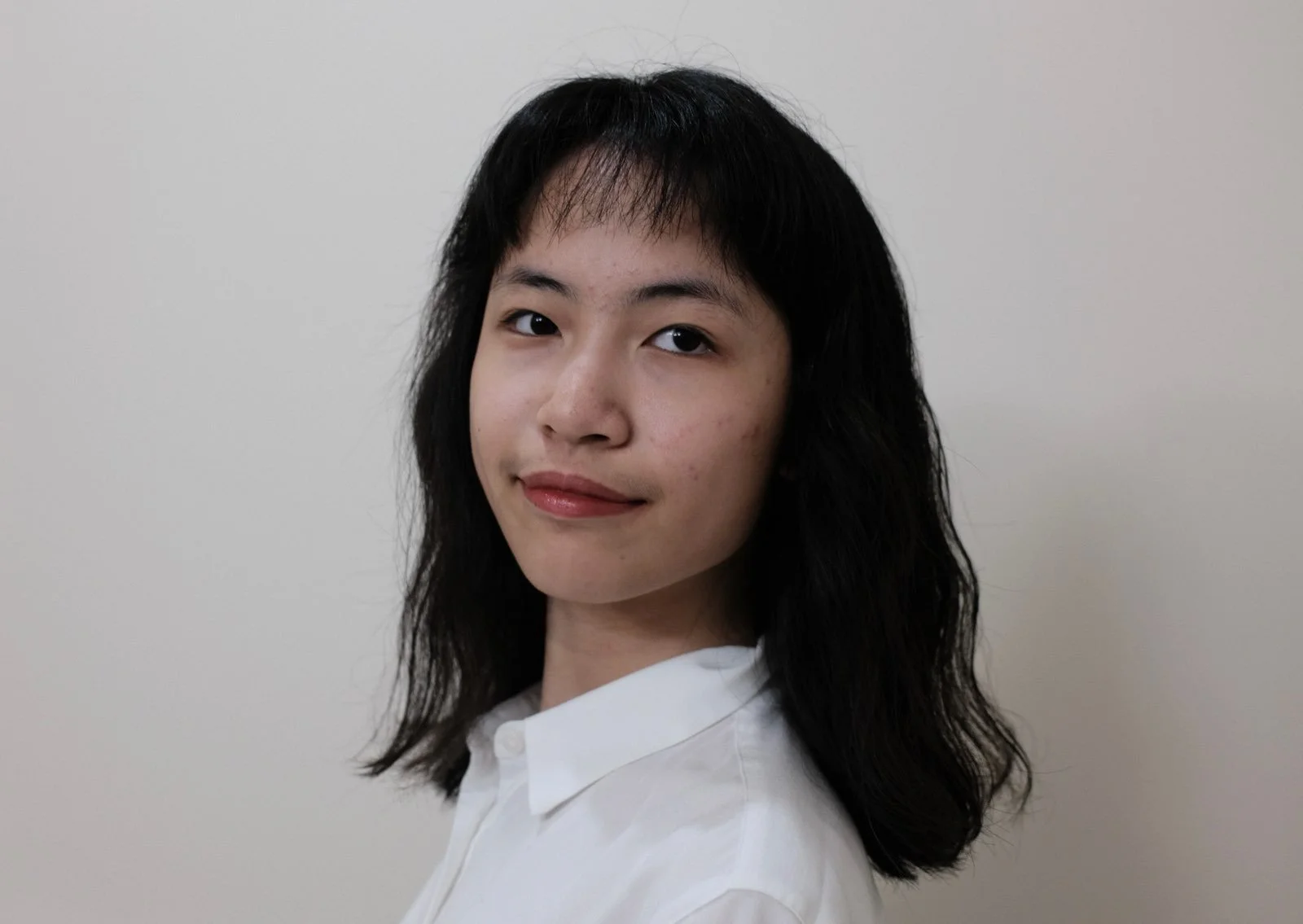 A young woman with medium-length dark hair and a white shirt looks at the camera with a slight smile against a plain light-colored background.