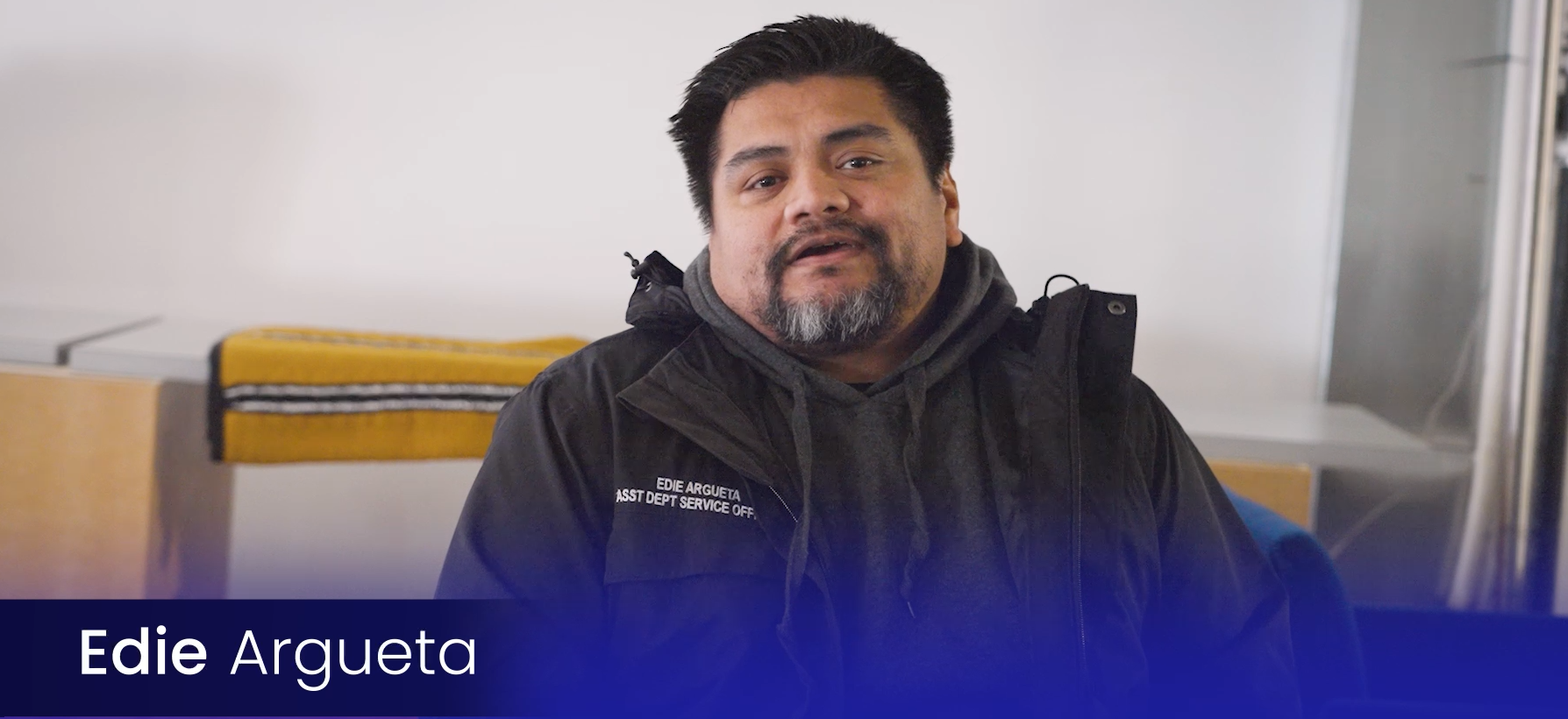 Man with dark hair, beard, and mustache wearing a black jacket and gray hoodie, sitting indoors in front of a white wall, with a yellow and black item and seating visible in the background, looking at the camera.
