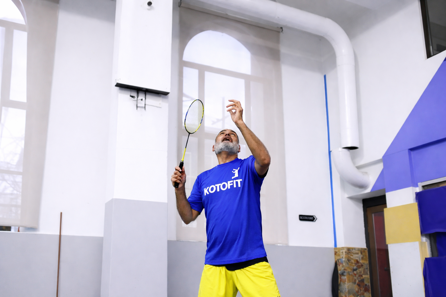 An older man with a gray beard playing indoor badminton, wearing a blue T-shirt and yellow shorts.