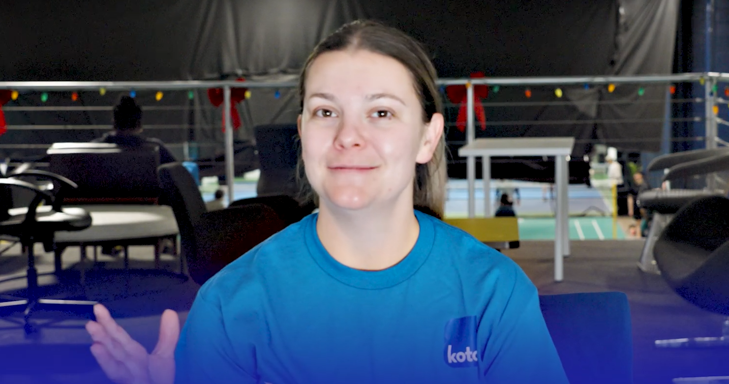 A young woman with brown hair wearing a blue shirt, sitting indoors in a setting with chairs and tables, possibly at a sports event or conference.
