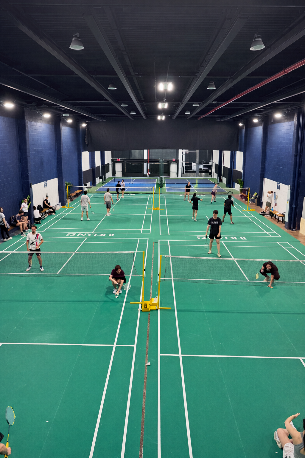 Indoor badminton court with multiple players playing, spectators sitting on benches, and court markings visible.