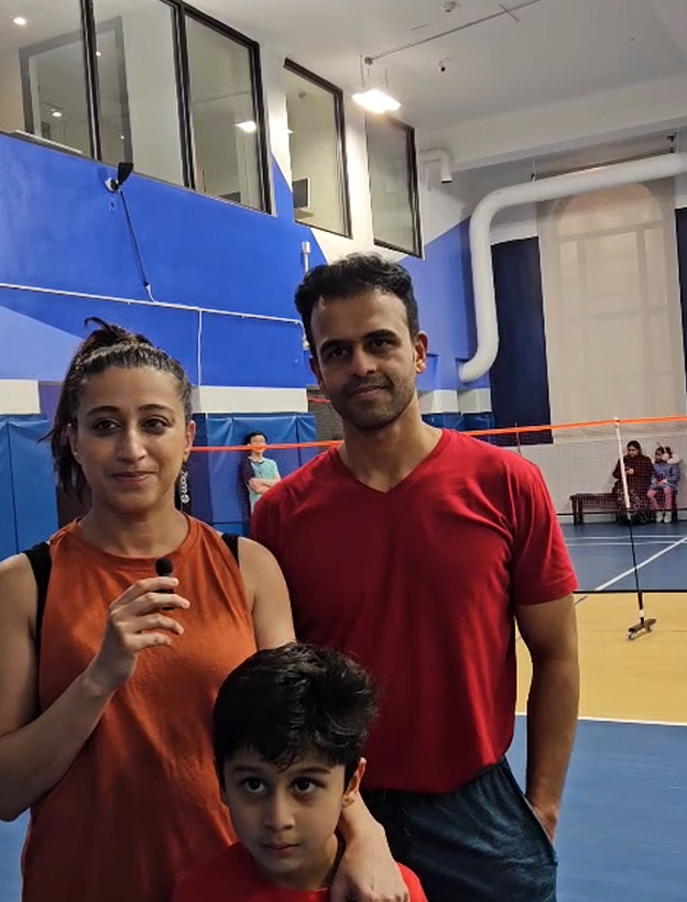 A woman, a man, and a young boy standing together in a gymnasium, with a badminton court and a net in the background.