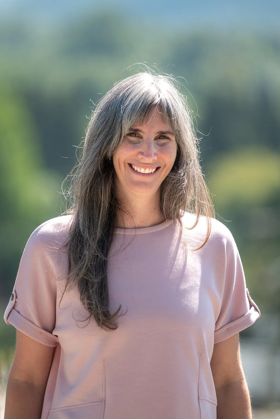 A woman with long gray hair smiling outdoors with a blurred green and blue background.