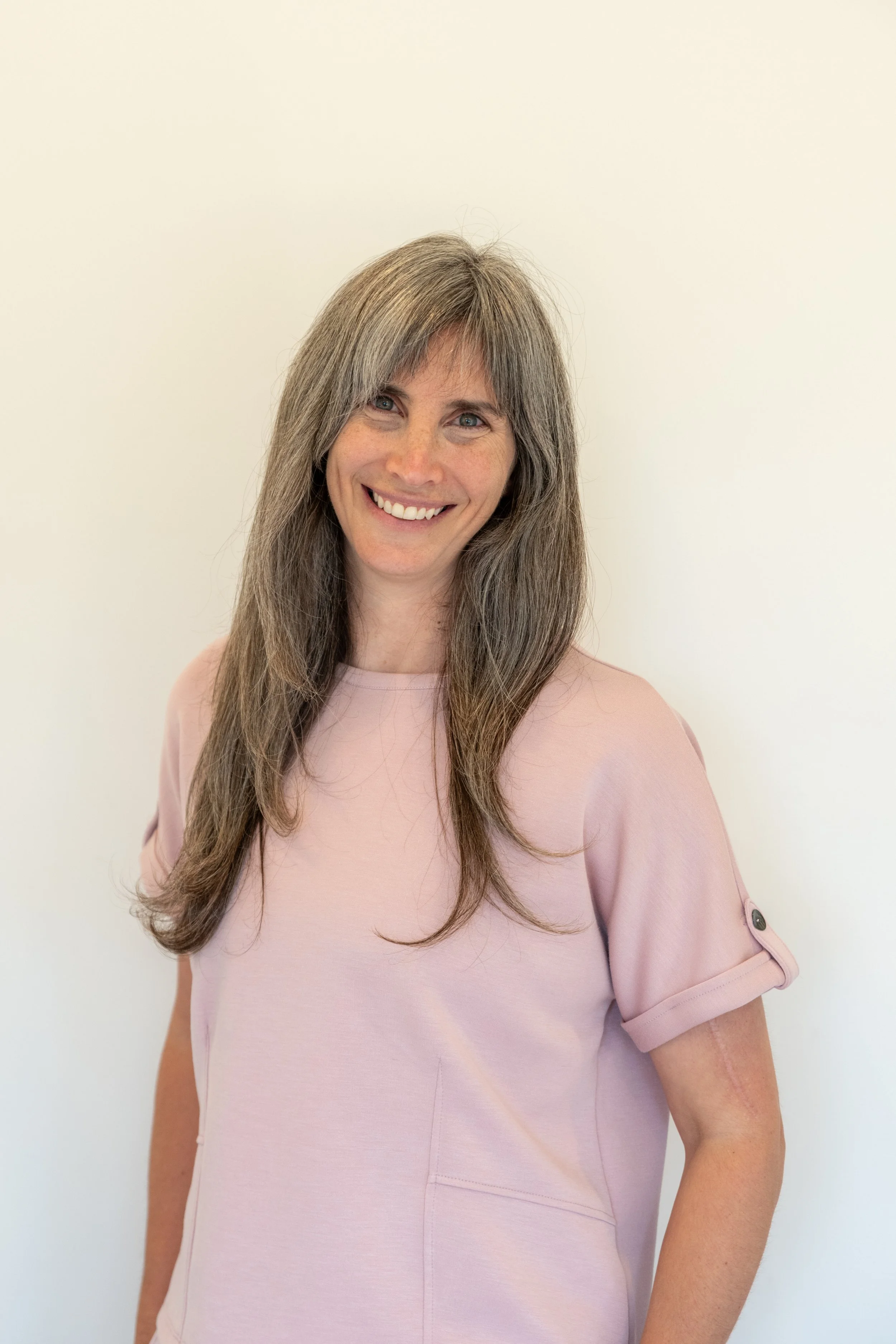 A woman with long gray hair, smiling, wearing a light pink top with rolled-up sleeves.