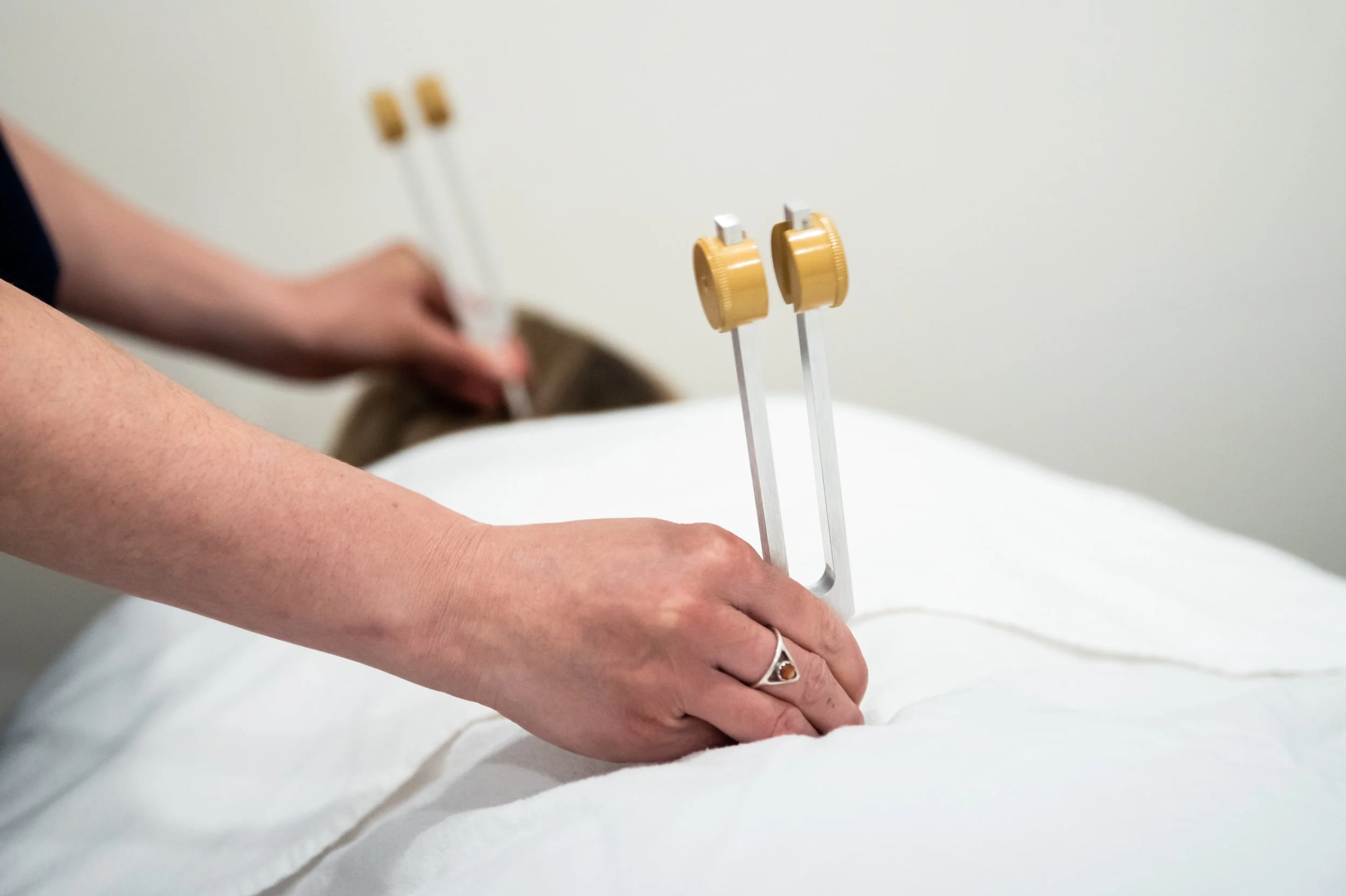 A person receiving a massage, with focus on their arm and hand resting on a white surface. Two gold-colored electrodes are attached to the person's forearm, connected to white wires for electrotherapy treatment.