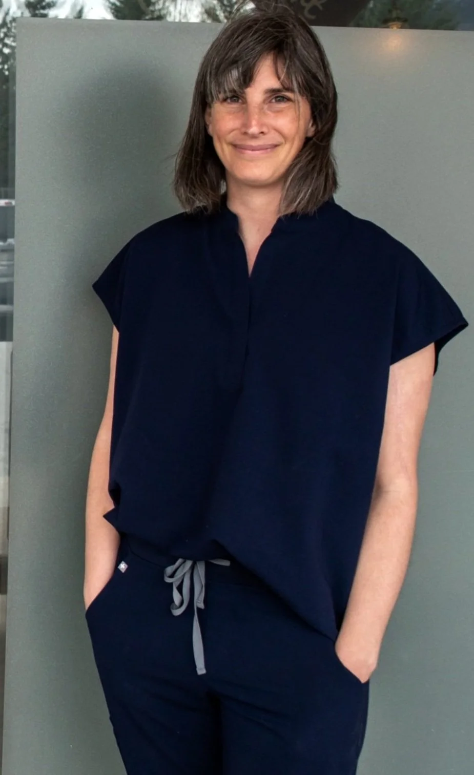A woman with shoulder-length brown hair and bangs, smiling, wearing a navy blue top and drawstring navy blue pants, standing outdoors against a gray background.