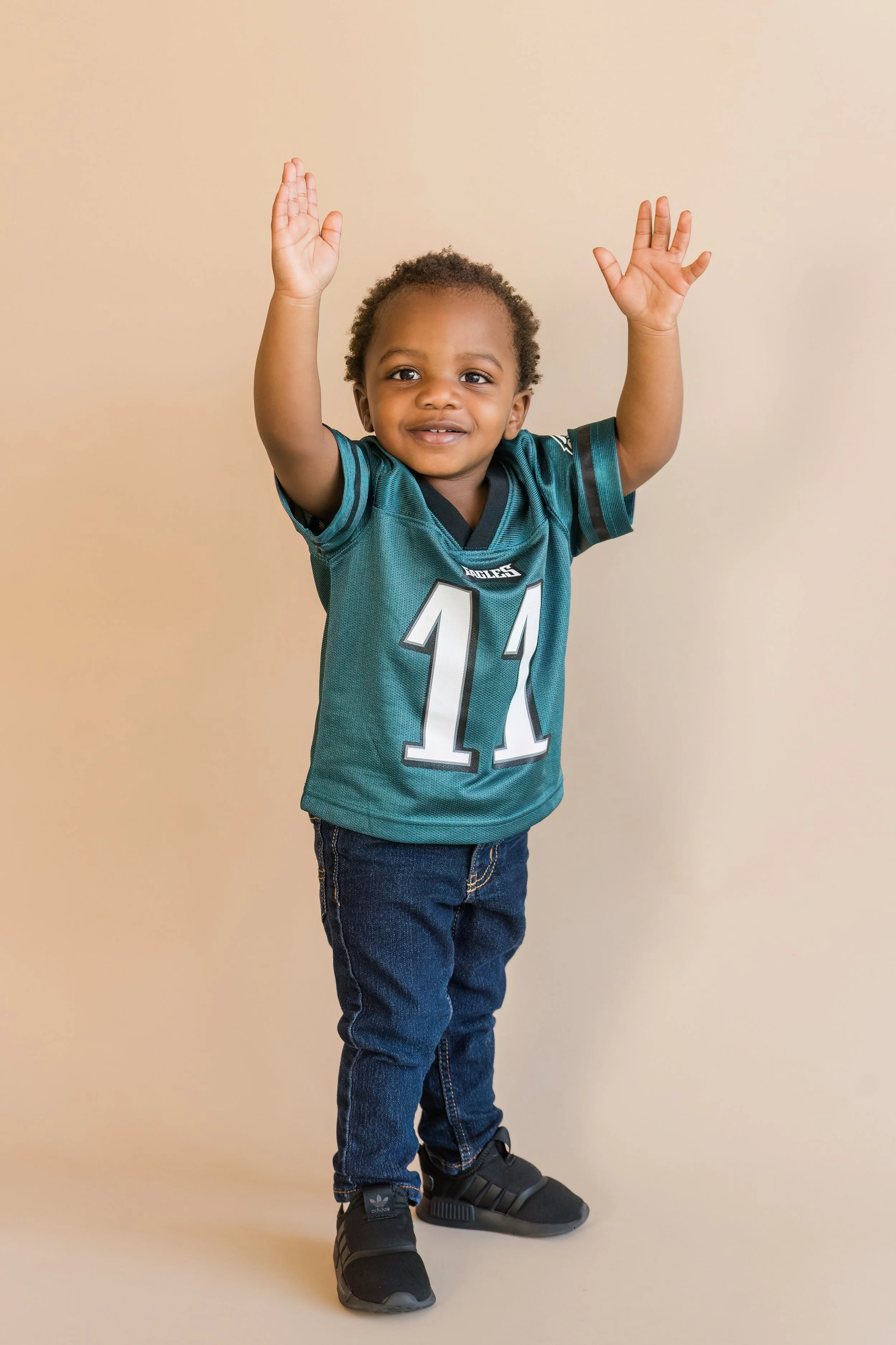 Young boy with curly hair wearing a teal football jersey with the number 11, jeans, and black sneakers, raising his arms and smiling.
