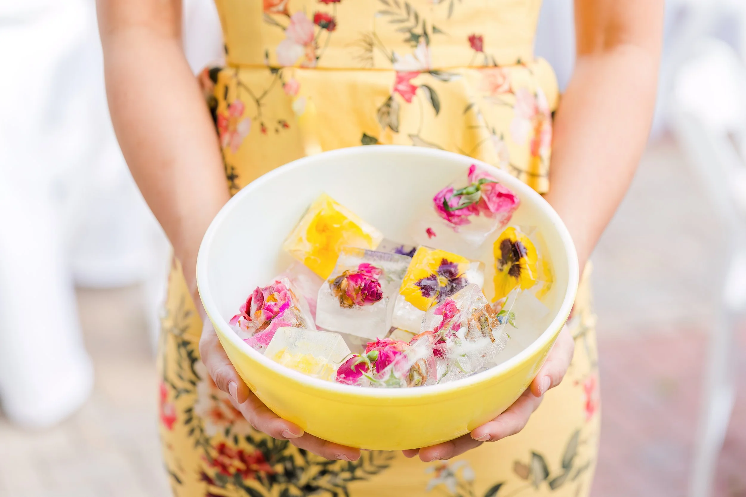 A person wearing a yellow floral dress holding a yellow bowl filled with ice cubes and decorative flowers and fruit.