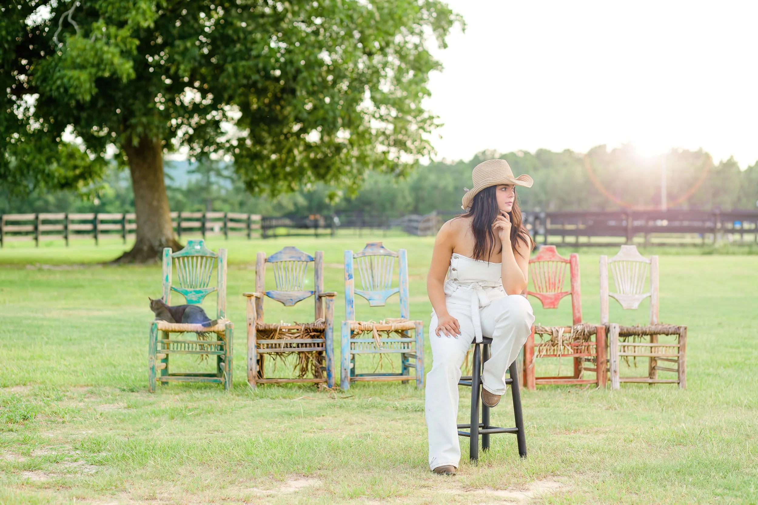 A woman with long dark hair sitting on a black stool in a field, wearing a wide-brimmed straw hat and white outfit, with a row of colorful rustic chairs behind her and a black cat resting on one of the chairs, under a large green tree during sunlight.