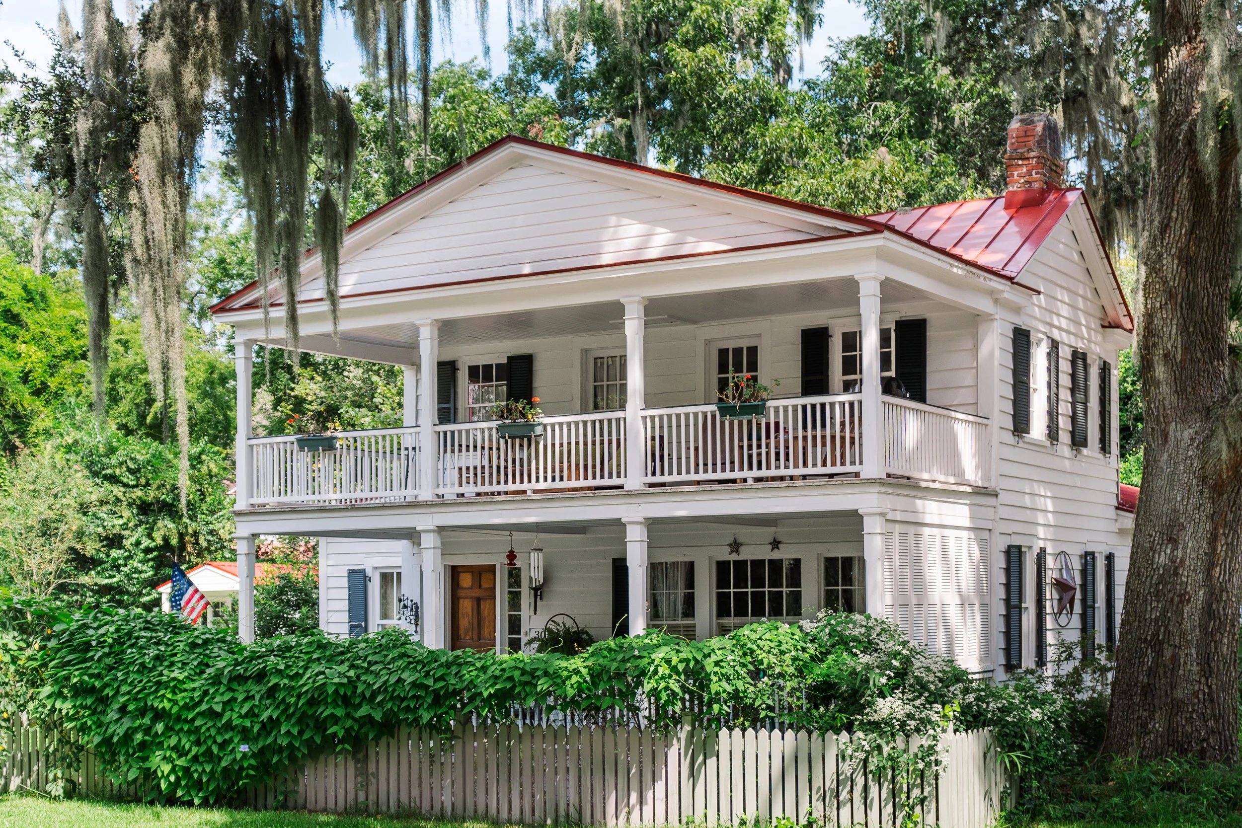A white two-story house with black shutters, a red metal roof, and a wraparound porch with chairs and hanging flower baskets. There's a fenced yard with greenery and tall trees surrounding the house.