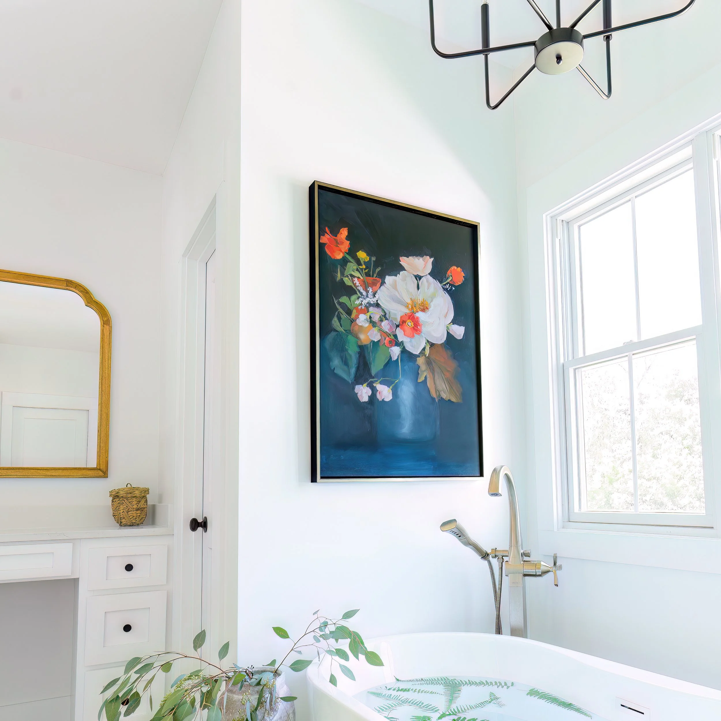 A bright bathroom with a large window, a black framed floral painting, a white bathtub with greenery, a gold-framed mirror, and a modern ceiling light fixture.