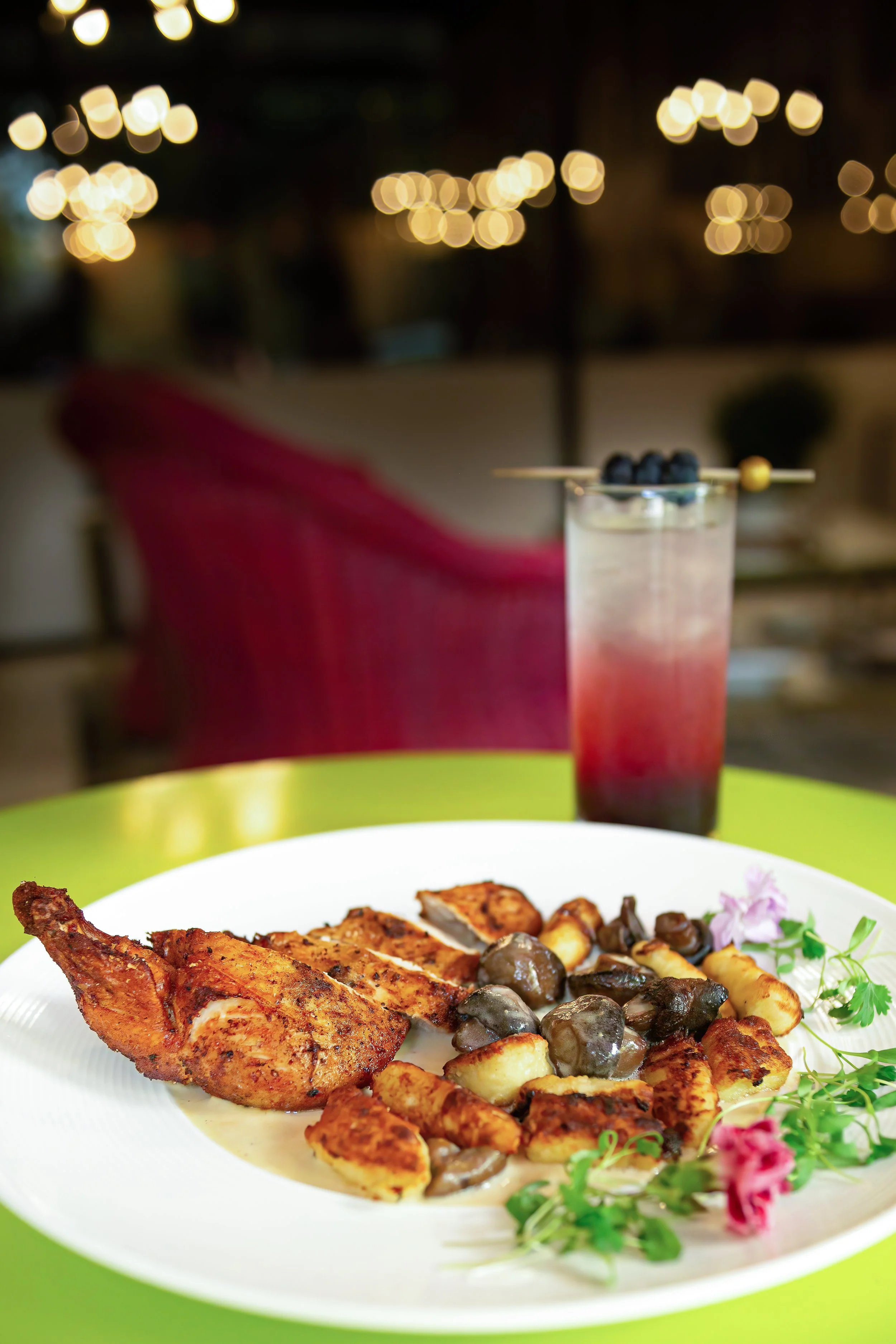 Plate of grilled chicken, roasted potatoes, and mushrooms with garnishes, with a blueberry cocktail in the background.
